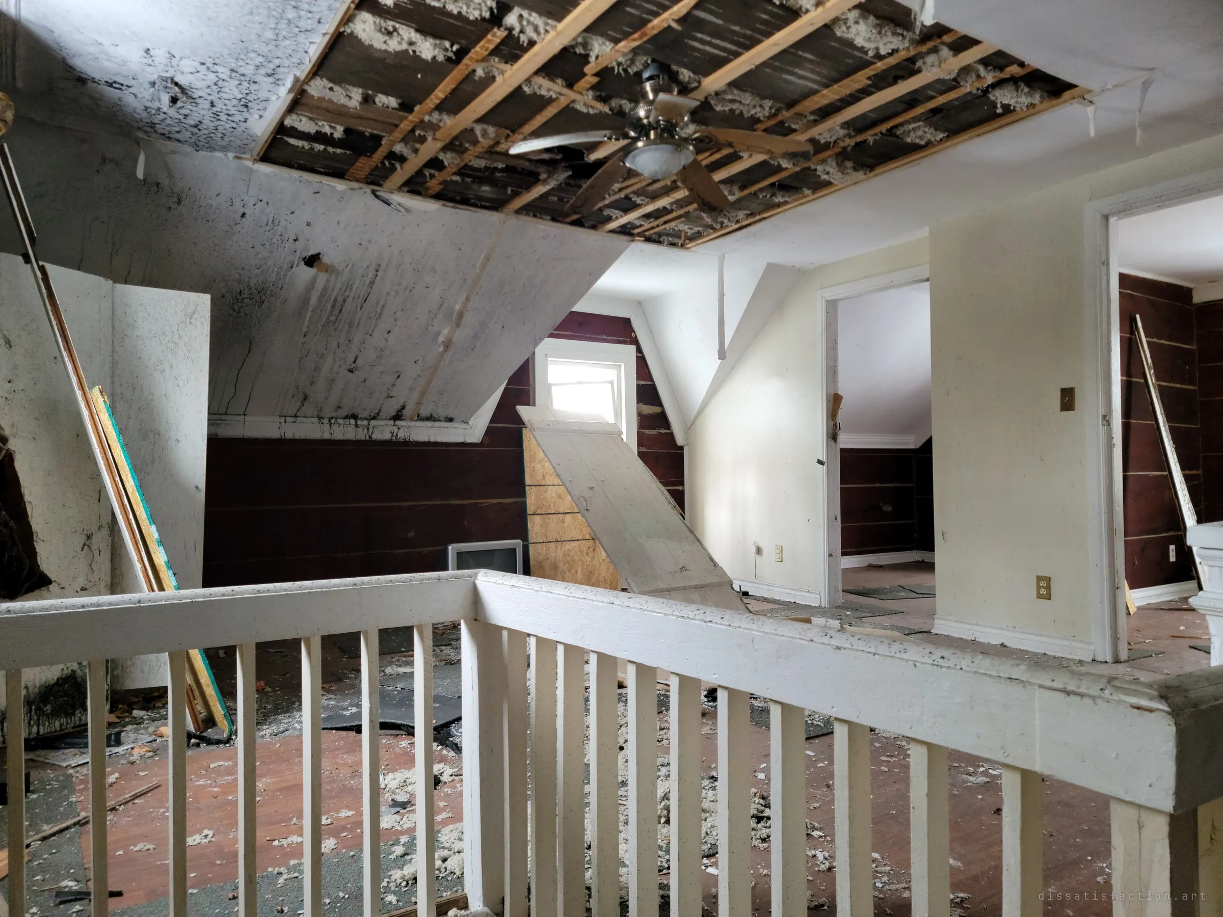 An upstairs landing behind a white railing, the ceiling torn open to expose lath and insulation around a hanging fan. Debris and loose boards cover the floor below.