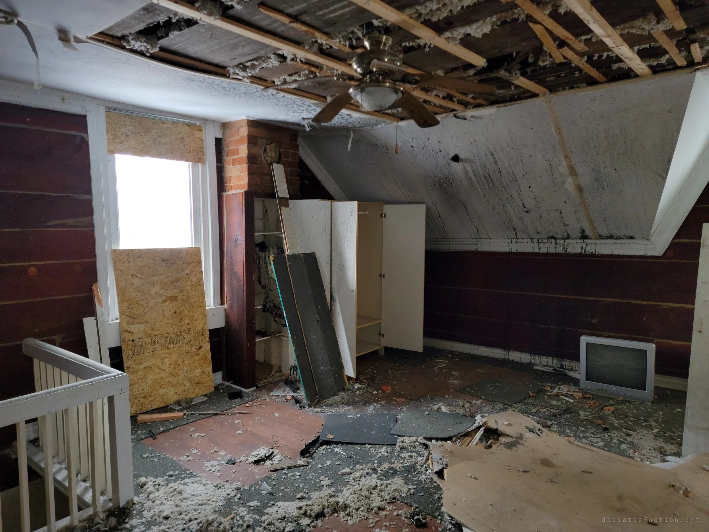 An attic bedroom with a gutted ceiling, a boarded window, an open wardrobe and an old CRT television amid insulation and scattered flooring tiles.