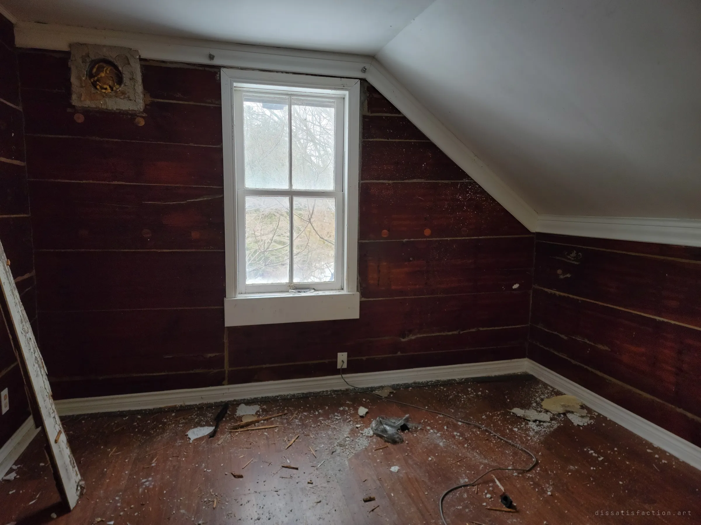 A small sloped-ceiling bedroom with dark red plank walls and a single window, its floor strewn with plaster chips and a stray cord.