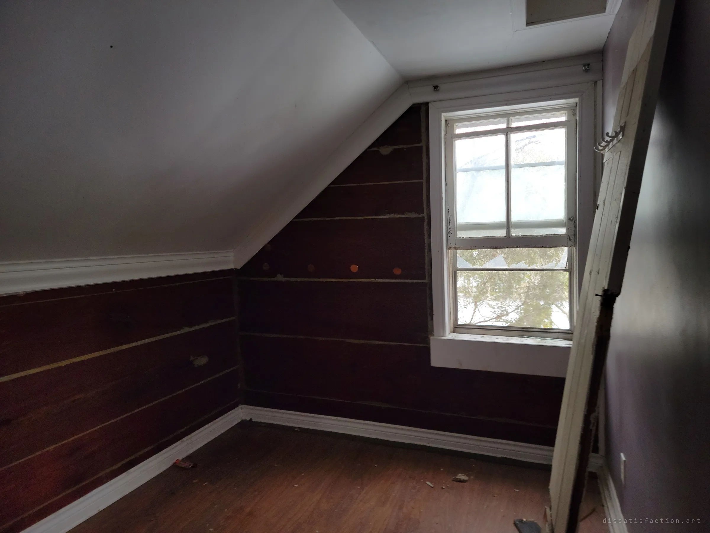 Another empty attic room under the slope of the roof, a loose door leaning against the wall beside a cracked window.