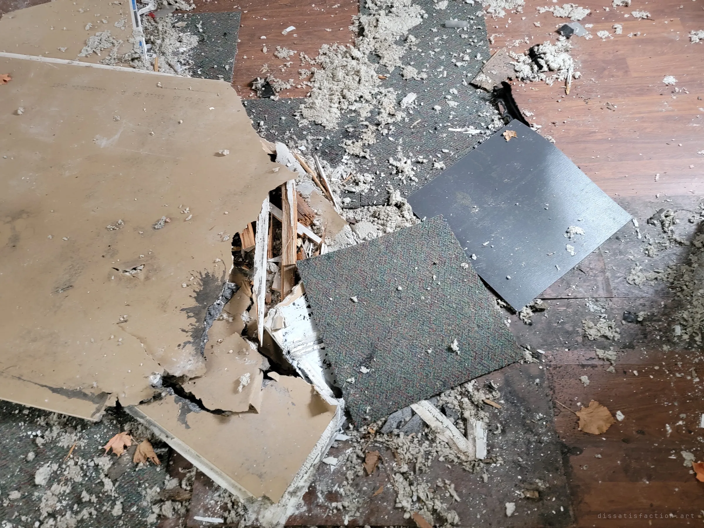 Close-up of the floor: a broken sheet of drywall punched through beside carpet tiles, with insulation and dead leaves everywhere.