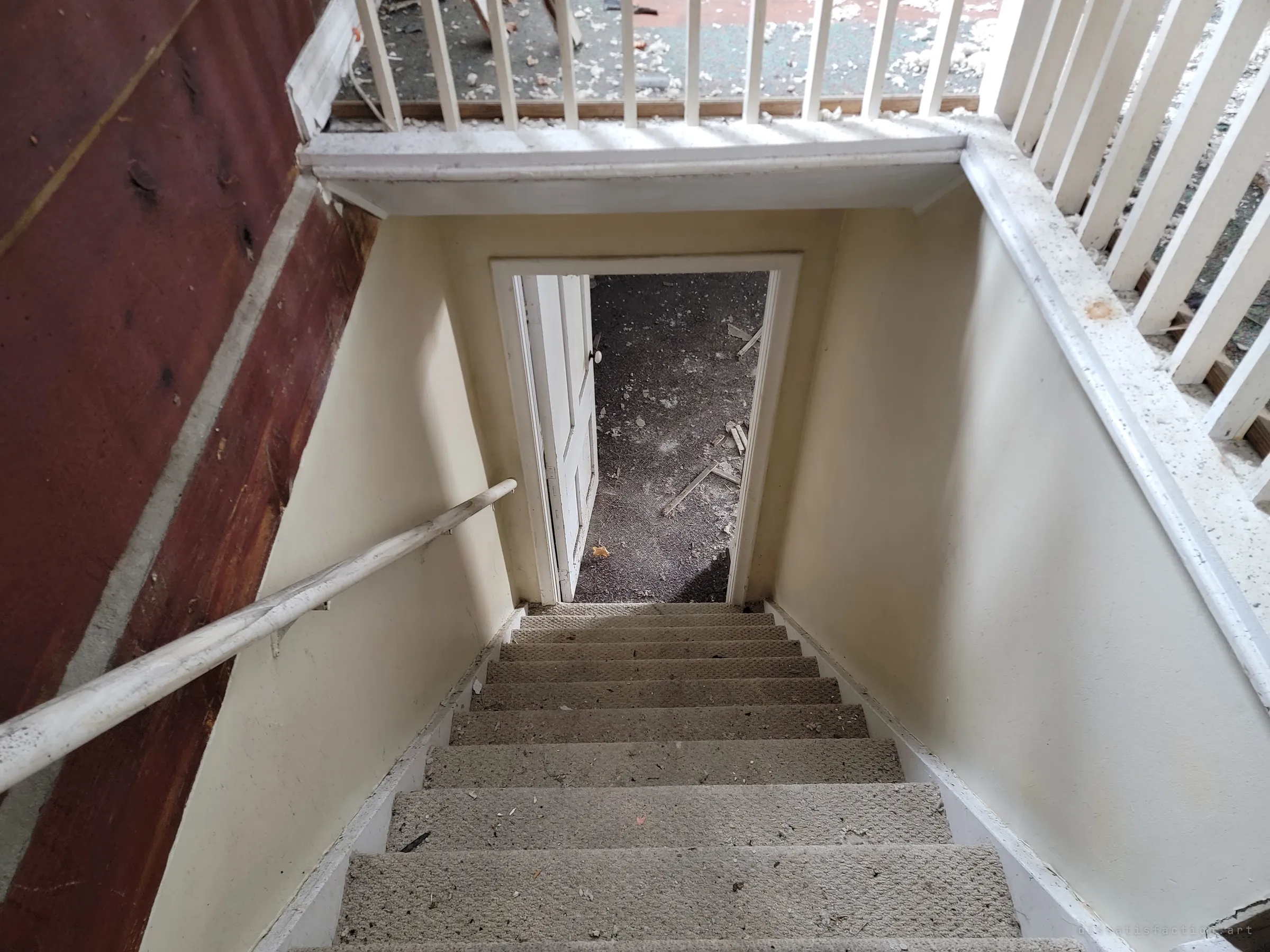 Looking down a long carpeted staircase from the upstairs railing to an open door and a debris-covered floor below.
