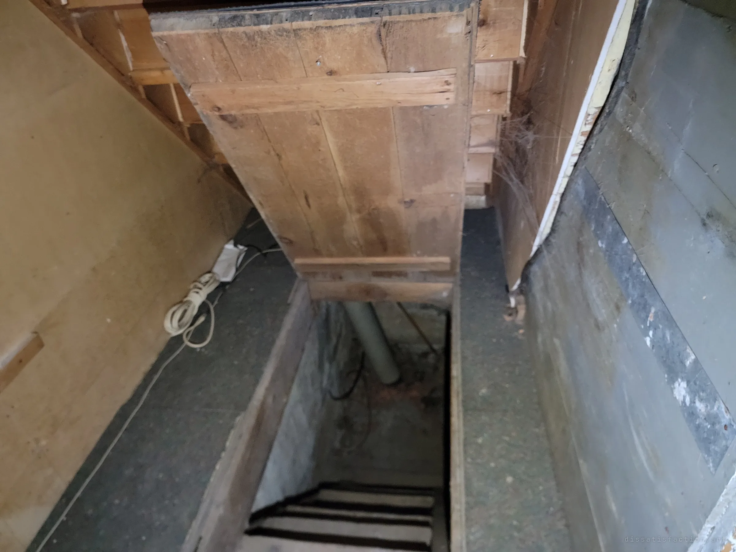 A tight wooden hatchway with a steep stair dropping into the dark basement, an extension cord coiled on the ledge.