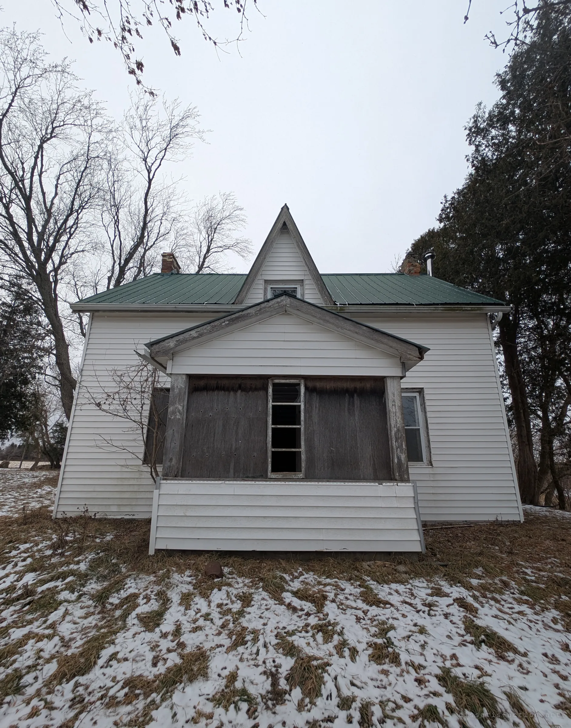 A white-sided farmhouse with a steep centre gable and green metal roof, its porch boarded over, standing in patchy snow under a grey sky. The first look at the house before going in.