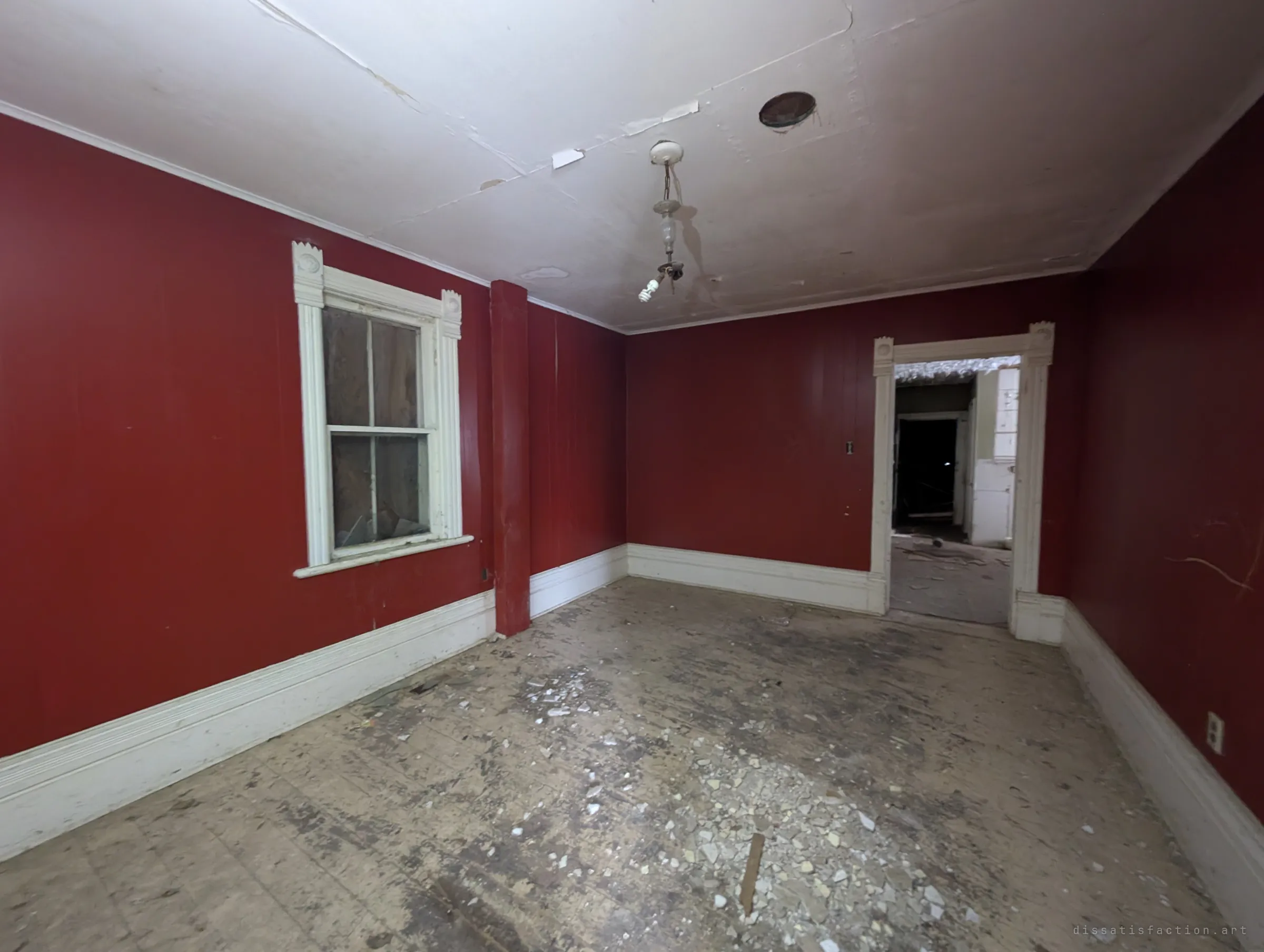 A deep red panelled room with white trim, a bare bulb hanging from the cracked ceiling and broken plaster scattered over the floor. A doorway leads into darker rooms beyond.