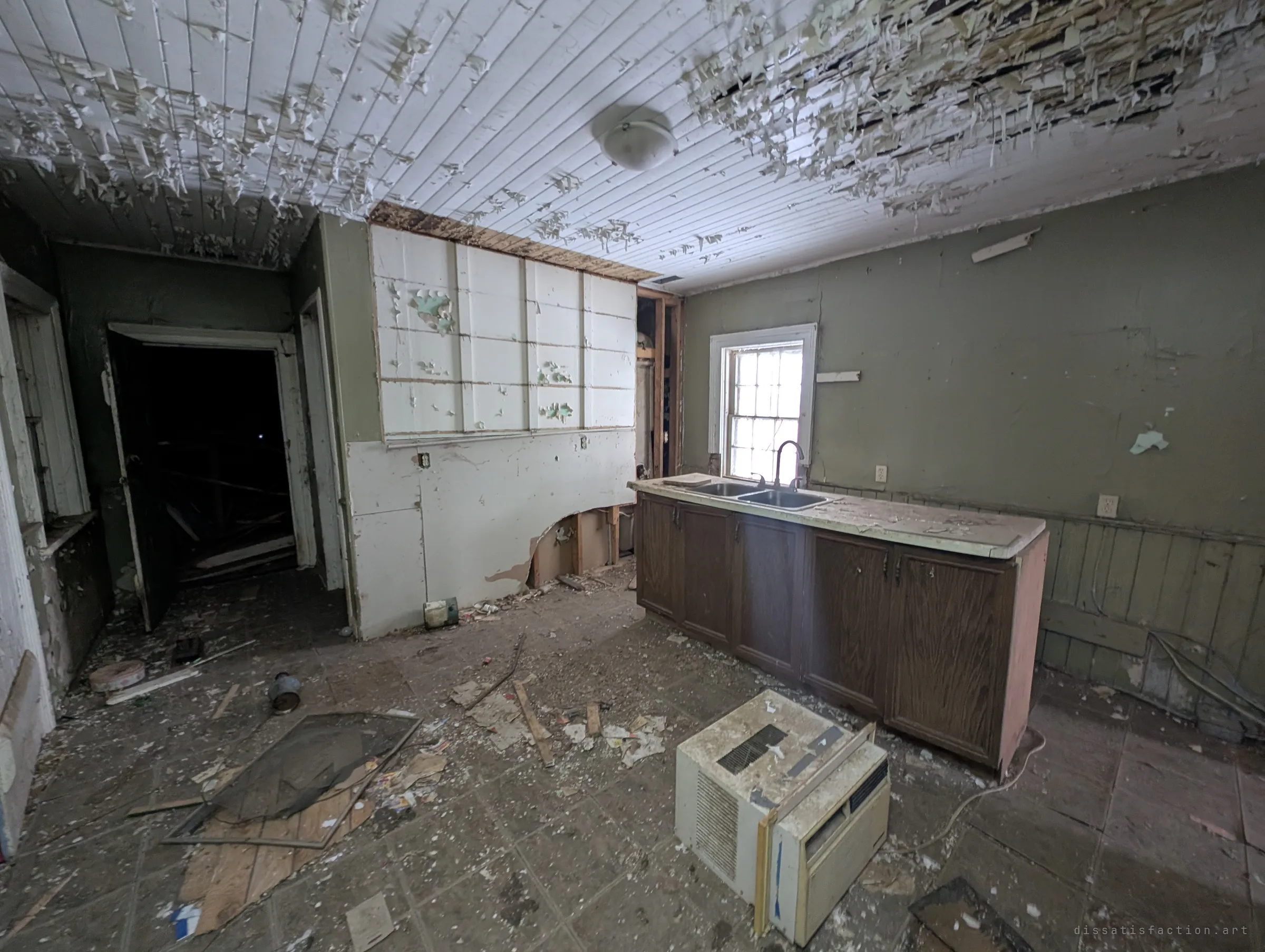 A gutted kitchen with paint peeling in curls from the plank ceiling, a lone sink cabinet under the window and a dumped window air conditioner on the littered floor.