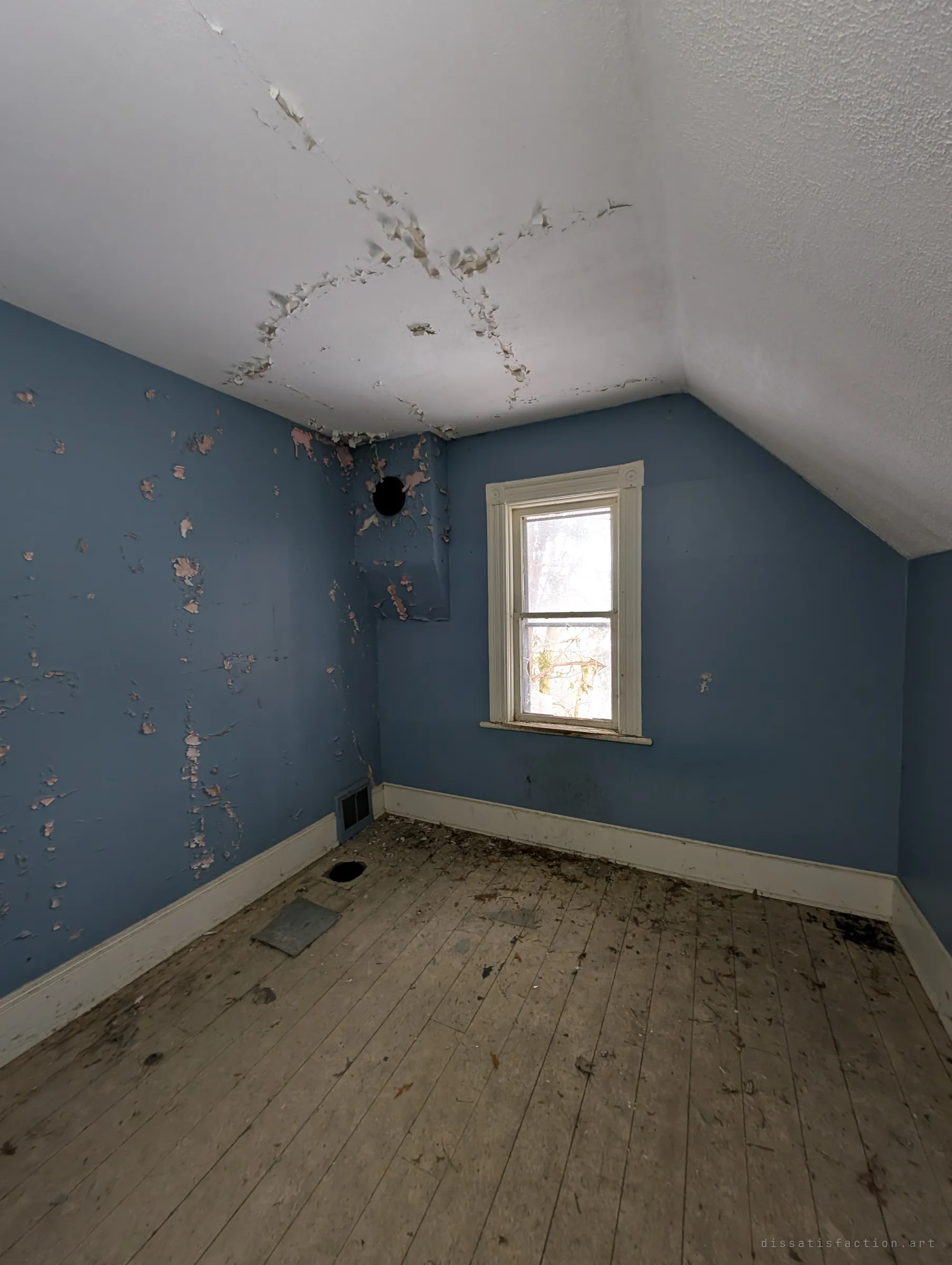 A small blue attic bedroom under a sloped ceiling, pink paint showing through where the blue has flaked away. A single window looks out onto bare branches.