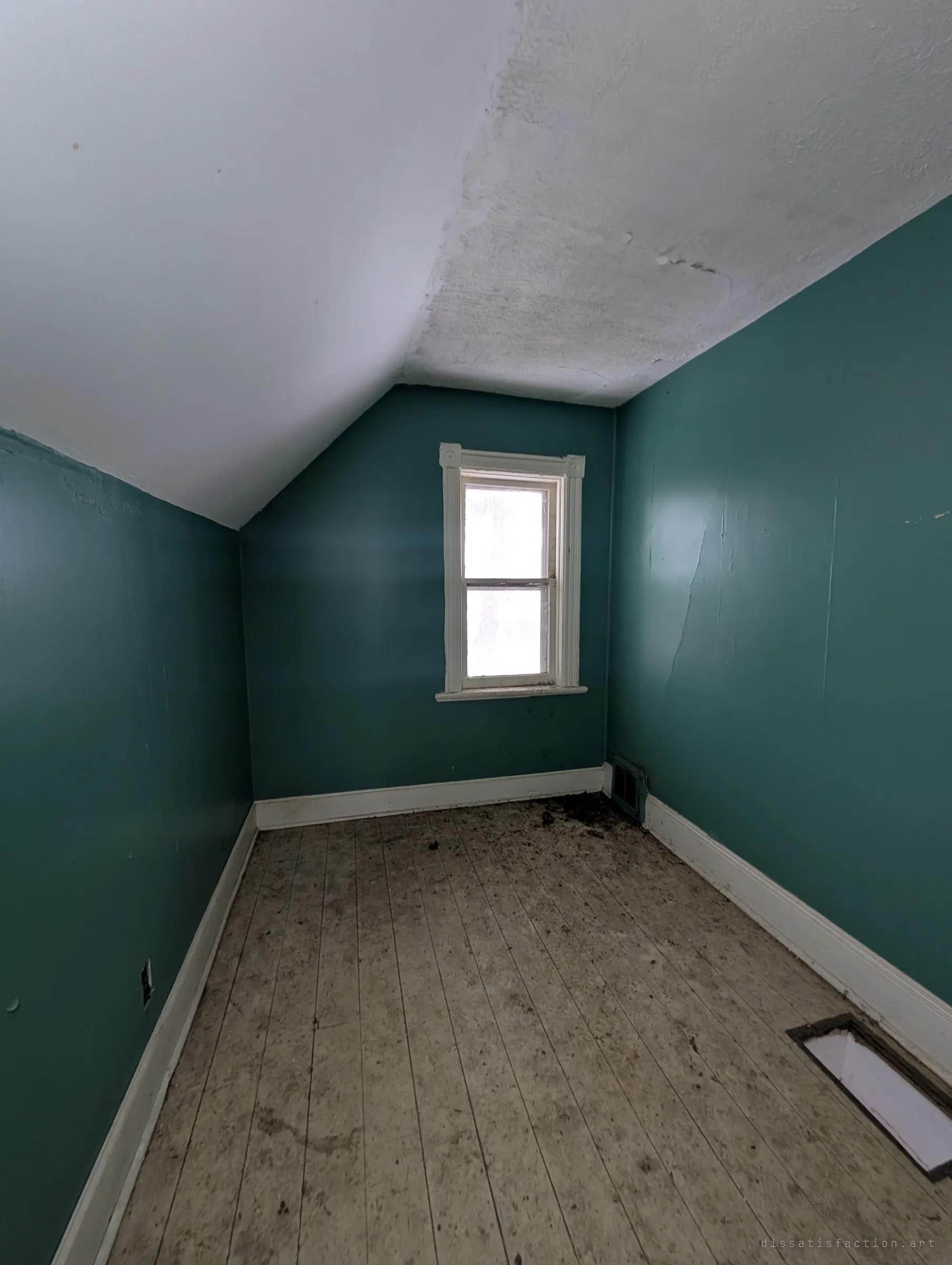A narrow teal room tucked under the roof slope, empty apart from dirt on the plank floor and an open floor vent. One small window lets in flat white light.