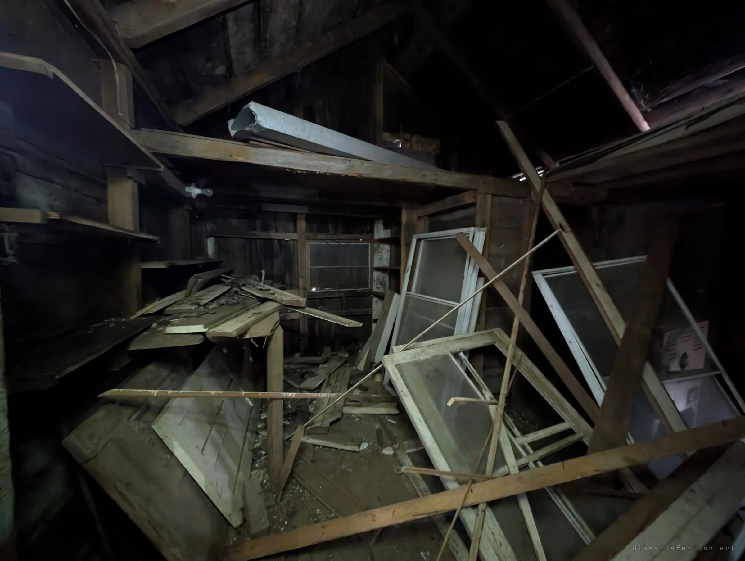 A dark wooden loft crammed with old window sashes, loose boards and fallen timbers leaning at every angle. Storage left to collapse on itself.
