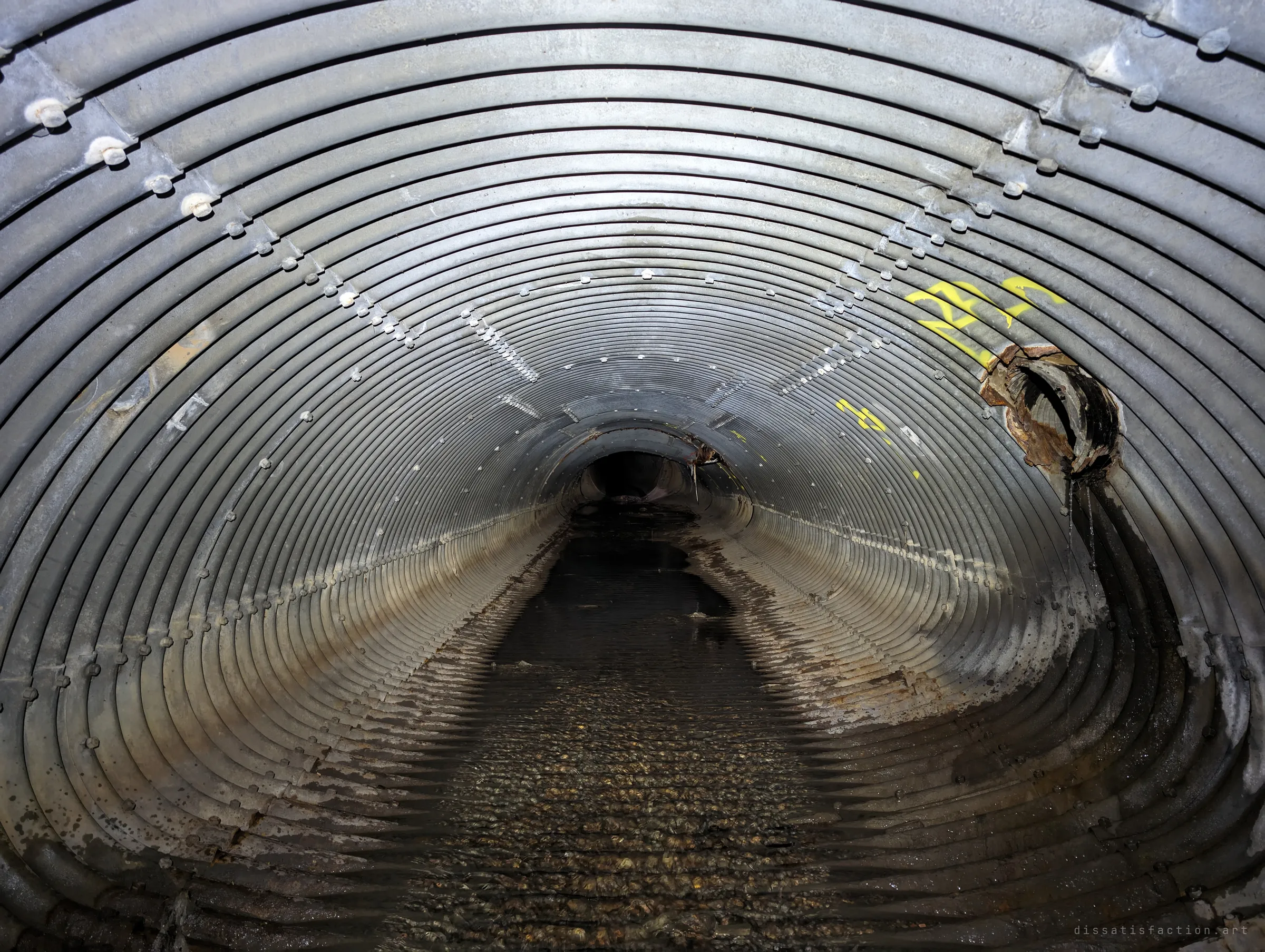 A long corrugated steel pipe recedes into darkness, with a rusted lateral pipe breaking through the wall on the right. Flashlight picks out the bolt rows and wet gravel floor.