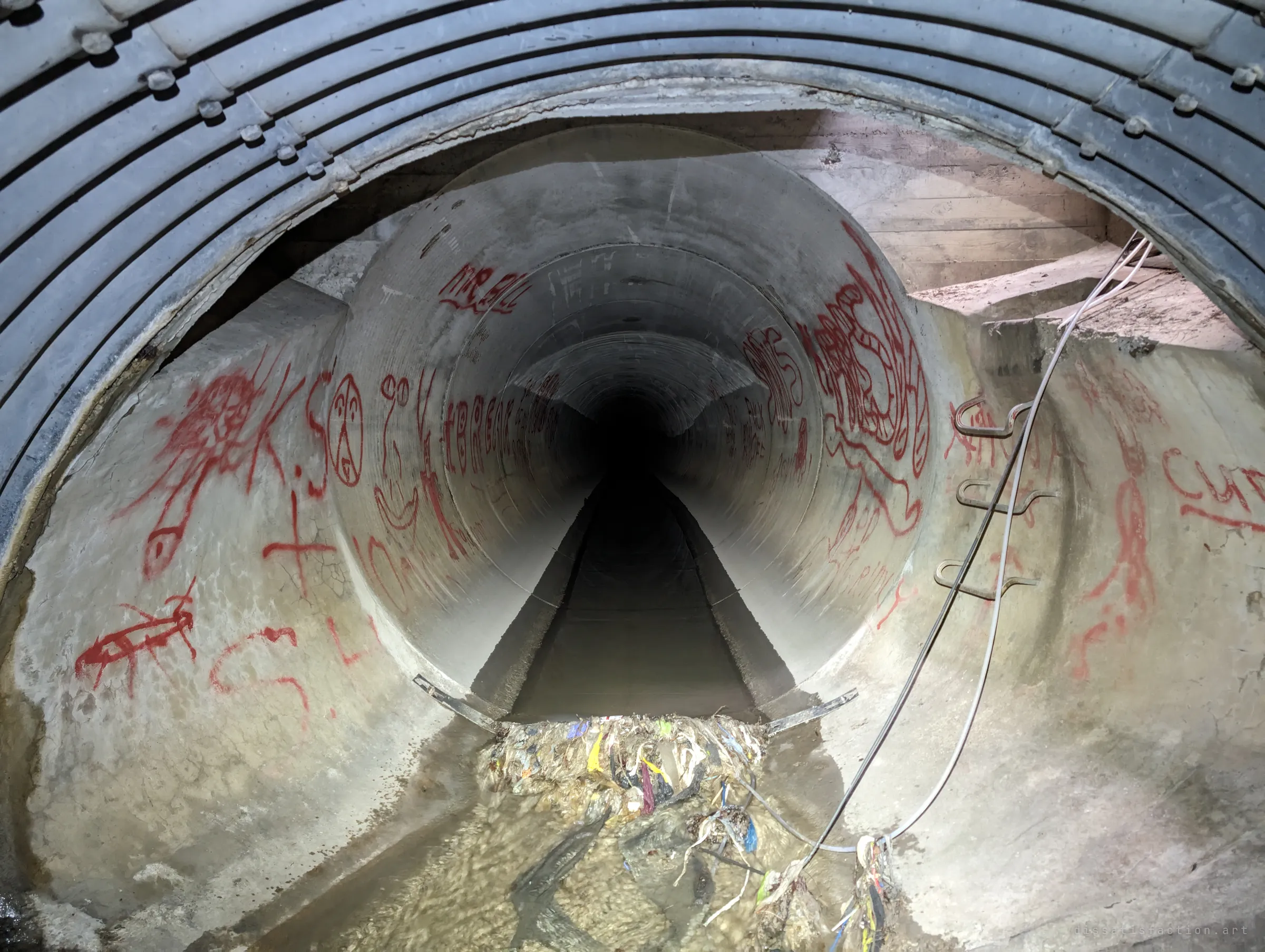 The corrugated pipe gives way to a smooth concrete pipe covered in red spray-painted scrawl, with step irons and a loose cable on the right. A debris mat has built up at the joint.