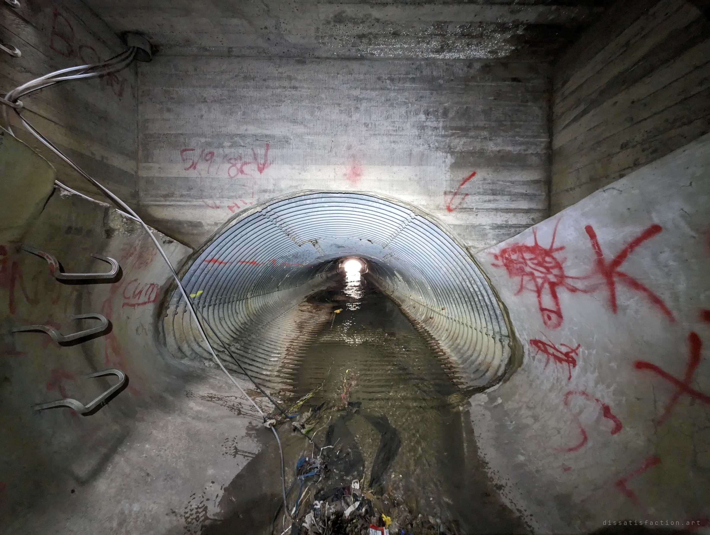 Looking back from the concrete chamber, the corrugated pipe runs toward a bright point of daylight. Red graffiti, step irons and a hanging cable line the chamber walls.