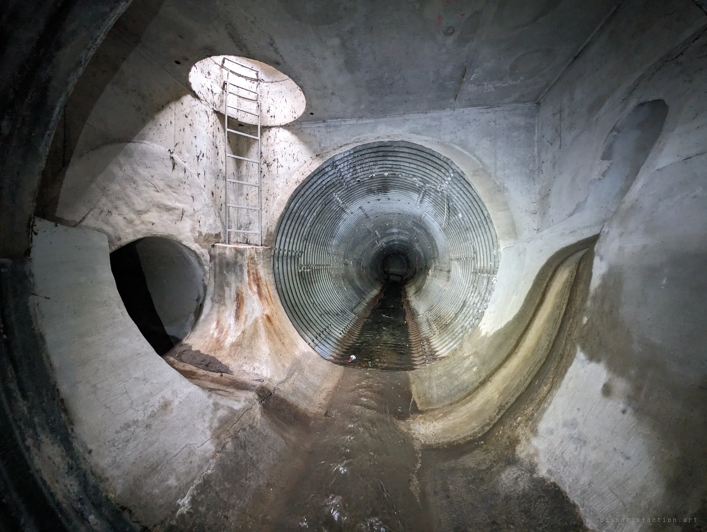 A concrete junction room where a corrugated pipe and a smaller side pipe meet, lit from above by a manhole shaft with a steel ladder. Curved concrete benching guides the flow toward the camera.