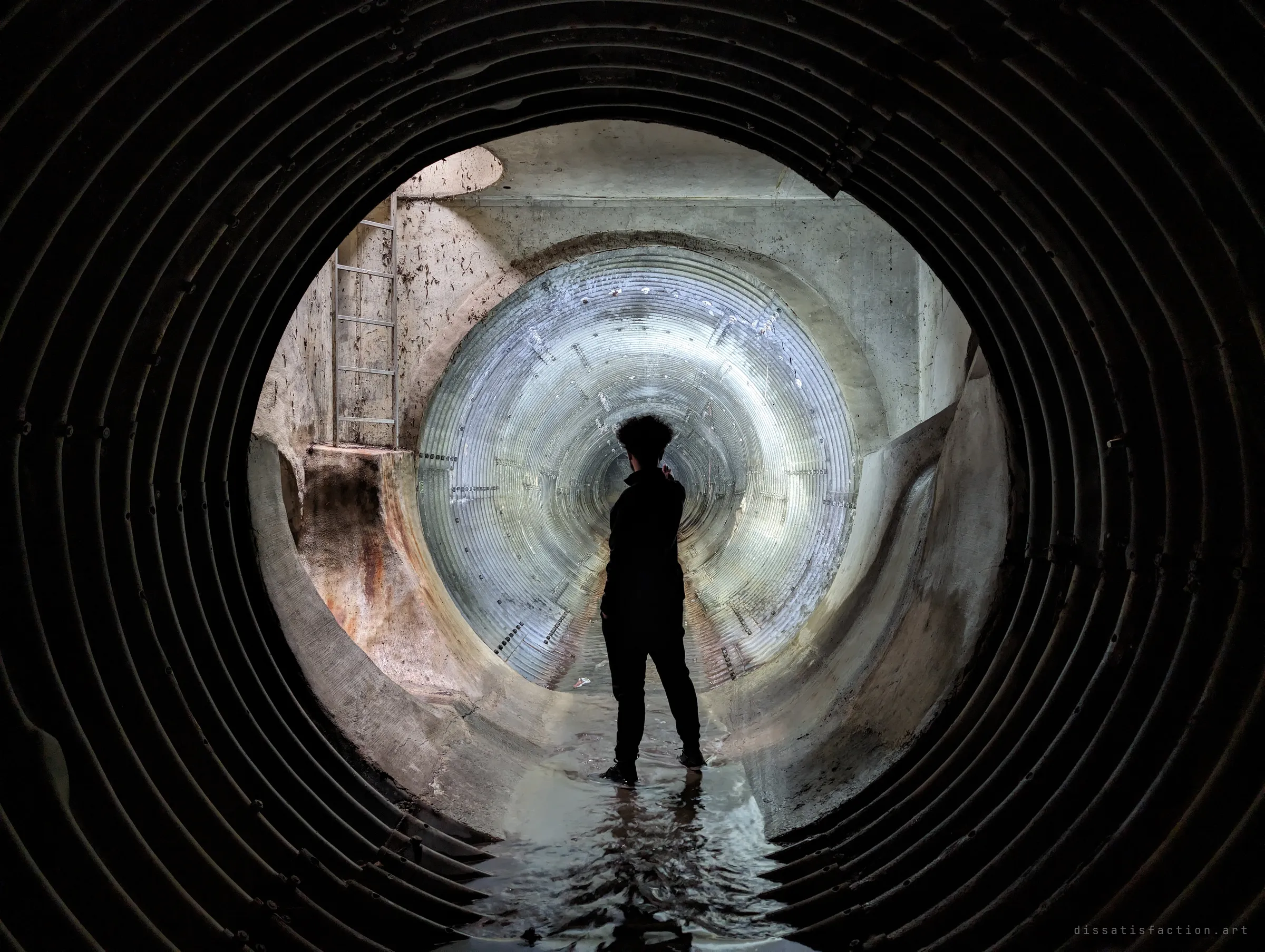 A silhouetted explorer stands in shallow water in the junction room, framed by the dark ring of the pipe and the lit corrugated tunnel behind. The figure gives the space its scale.