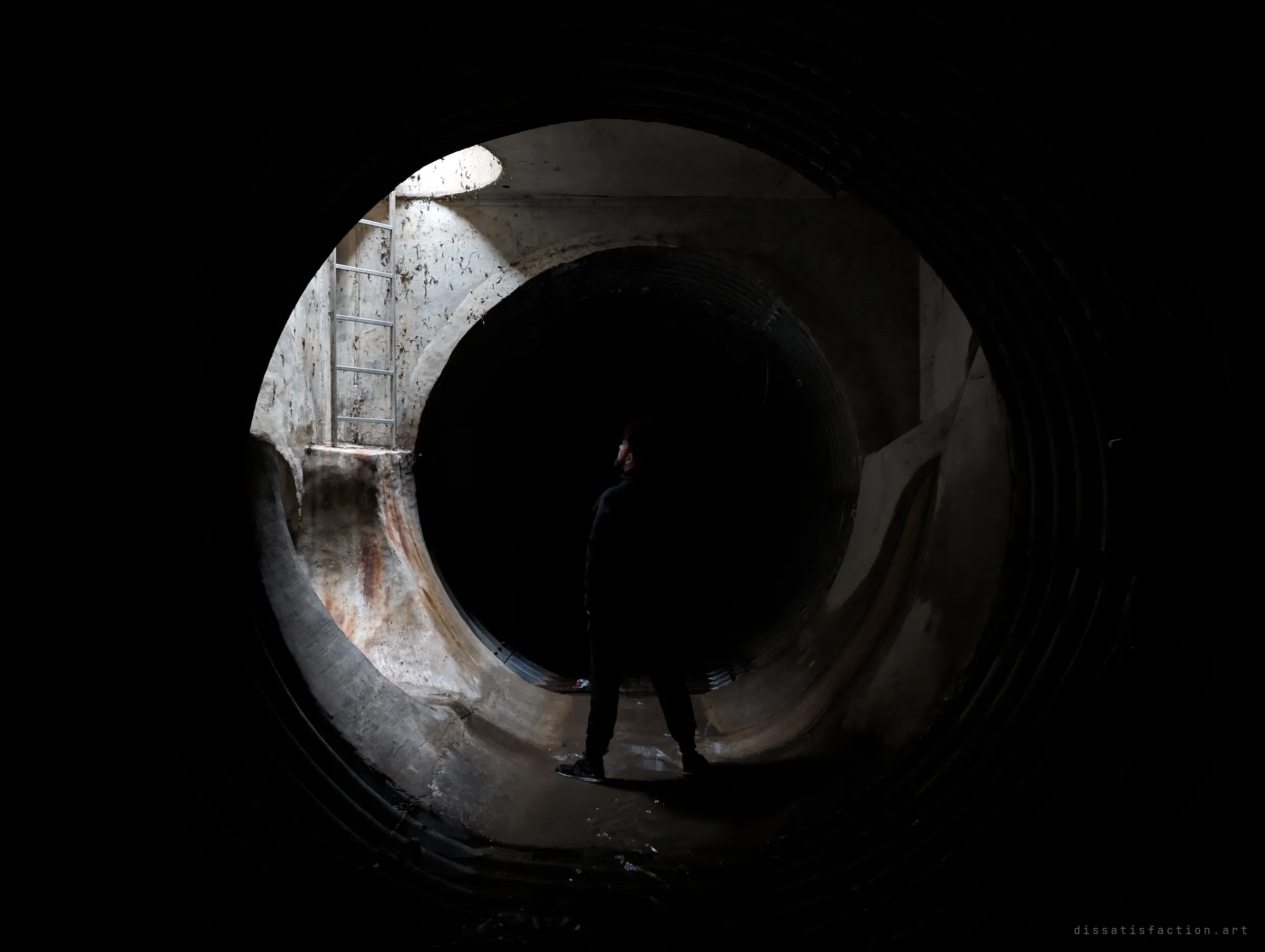 The same junction with the lights cut: only the manhole shaft's daylight catches the ladder and benching, the explorer nearly lost in shadow. A darker companion to the previous frame.