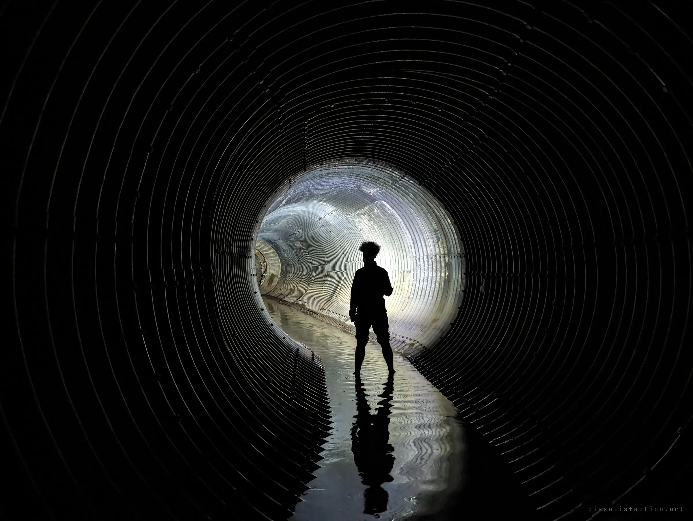 A lone silhouetted figure stands in shallow water inside a dark corrugated steel pipe, backlit by the bright bend of tunnel beyond. The ribs of the pipe frame the figure like rings of a target.