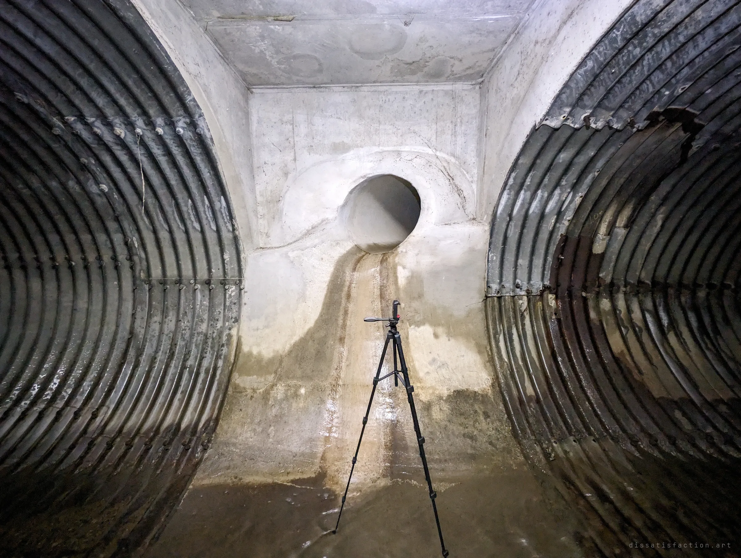 A concrete junction chamber between two corrugated steel pipes, with a smaller round inlet high on the back wall and a stained trail where water has run down. A tripod stands in the middle for scale.
