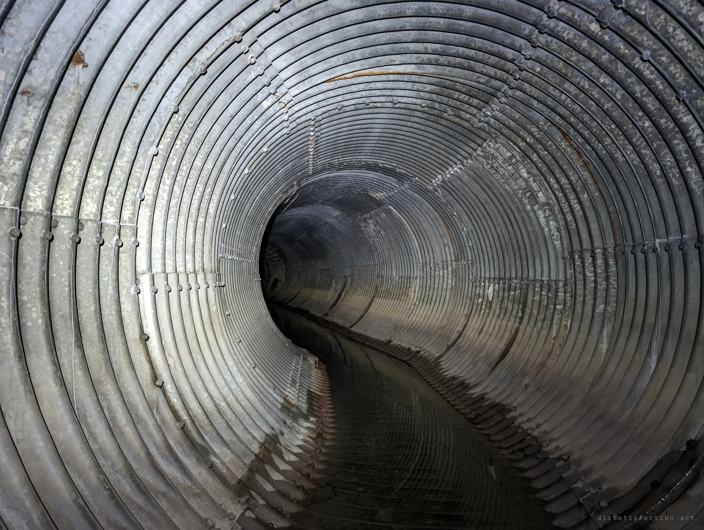 Looking down a long, gently curving corrugated steel pipe, its bolted seams and ribs spiralling into darkness. A thin sheen of water runs along the invert.