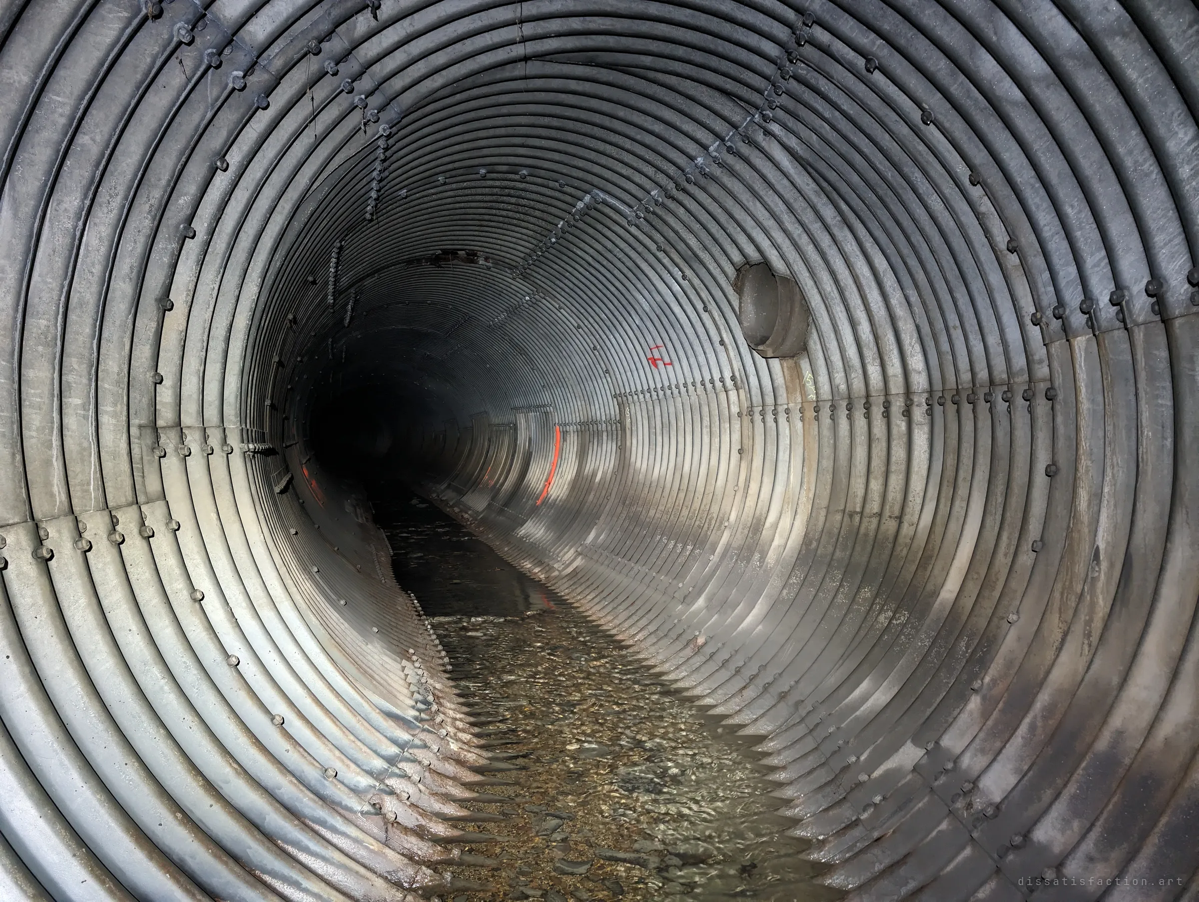A straight run of corrugated steel pipe with a capped side connection and faded orange survey paint on the wall. Clear water flows over gravel along the bottom.