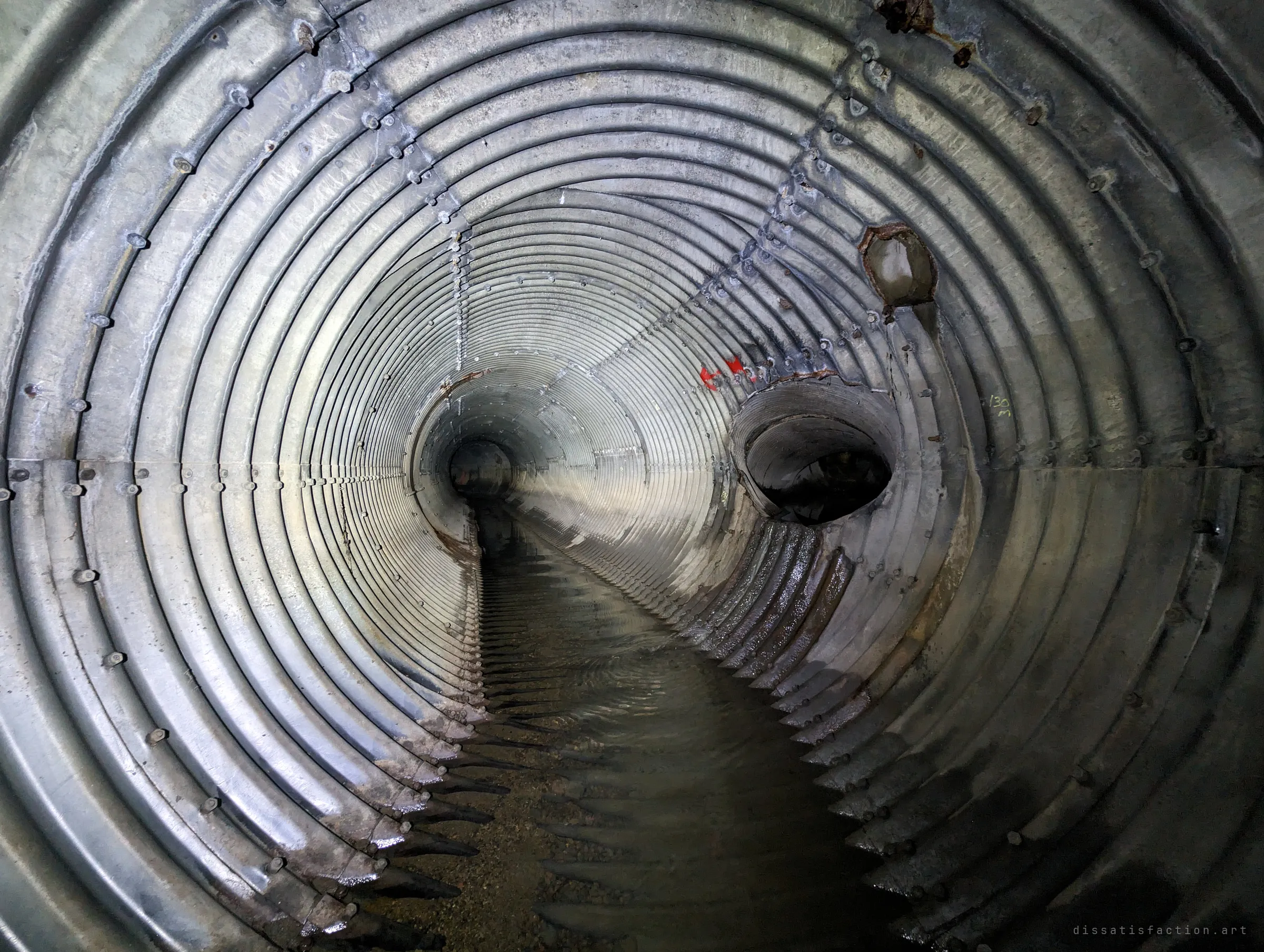 Two side pipes break into the main corrugated tunnel, one ragged and rusted at the edge, beside red paint arrows and a hand-written distance marker. The pipe narrows ahead into a smaller section.