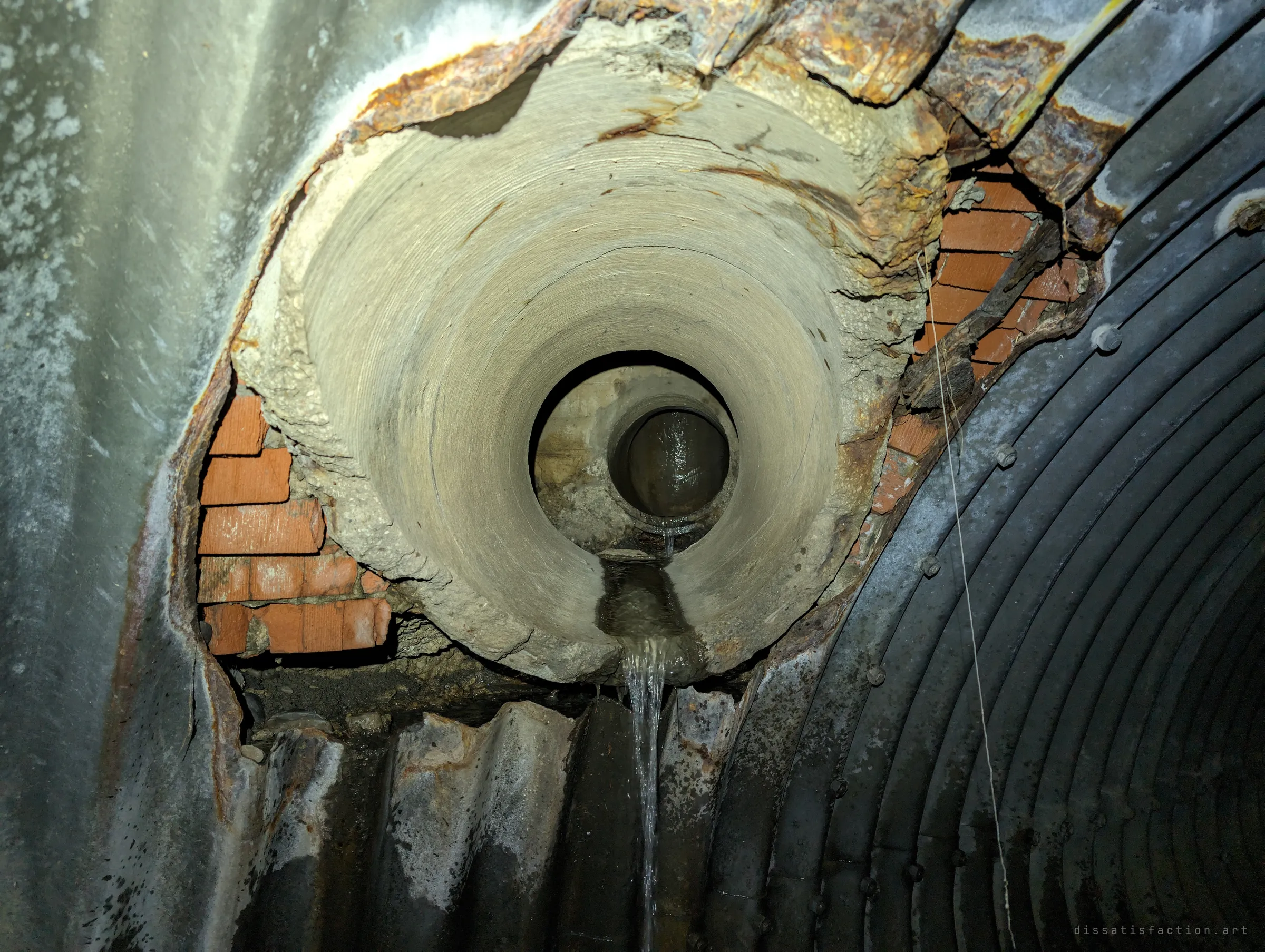 A crumbling concrete inlet punched through rusted corrugated steel, with old red brick exposed around it and a thin stream of water pouring out of a smaller pipe nested inside. It shows how badly the structure has decayed.