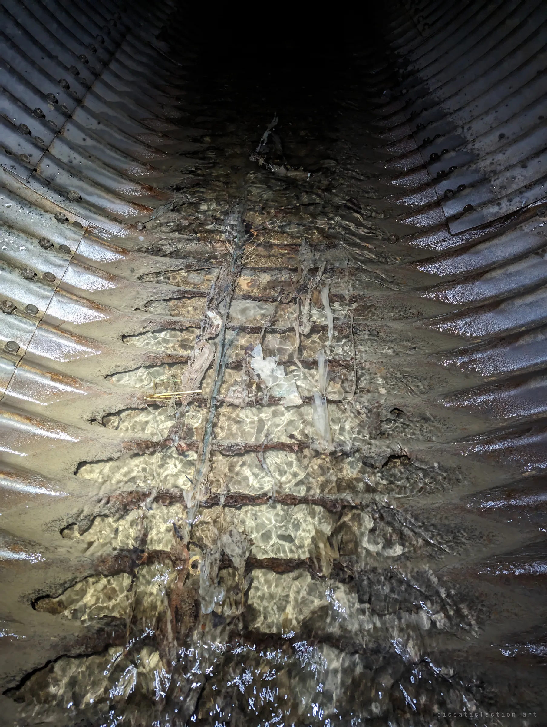 Water ripples over the bottom of a corrugated pipe where the steel has rotted away, leaving only rusted ribs and bare ground beneath. The corrosion runs down the centre of the pipe into the dark.