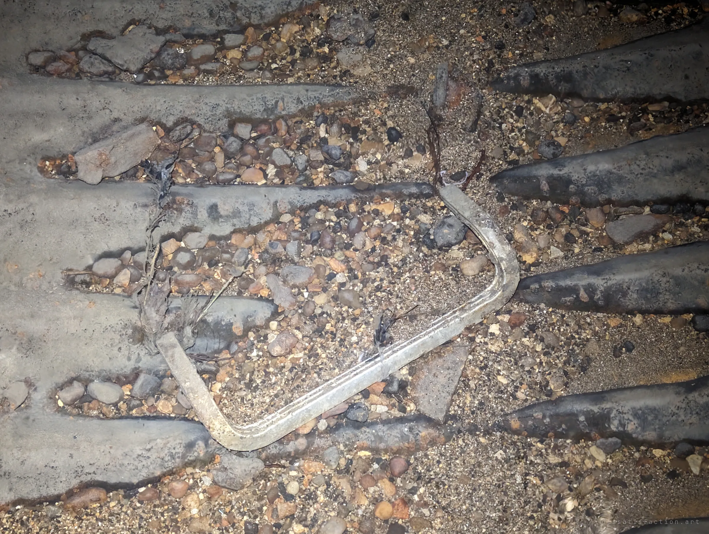Close-up of the pipe floor: rusted corrugation ridges half buried in gravel and pebbles, with a bent metal bracket lying among them. A small detail of the debris the water leaves behind.