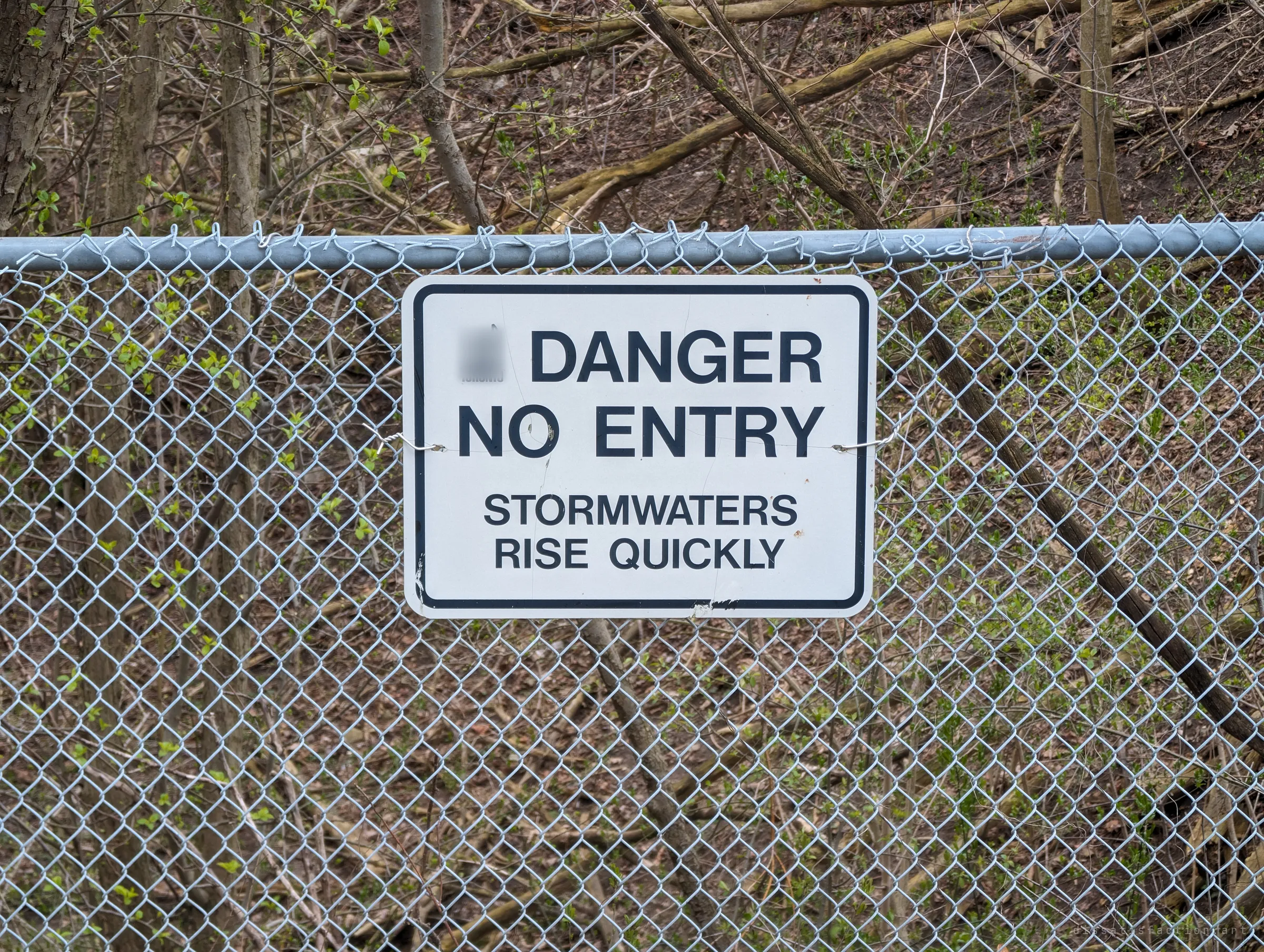 A danger, no entry sign warning that stormwaters rise quickly, wired to a chain-link fence in front of a bare spring ravine. It marks the way in to the drain.