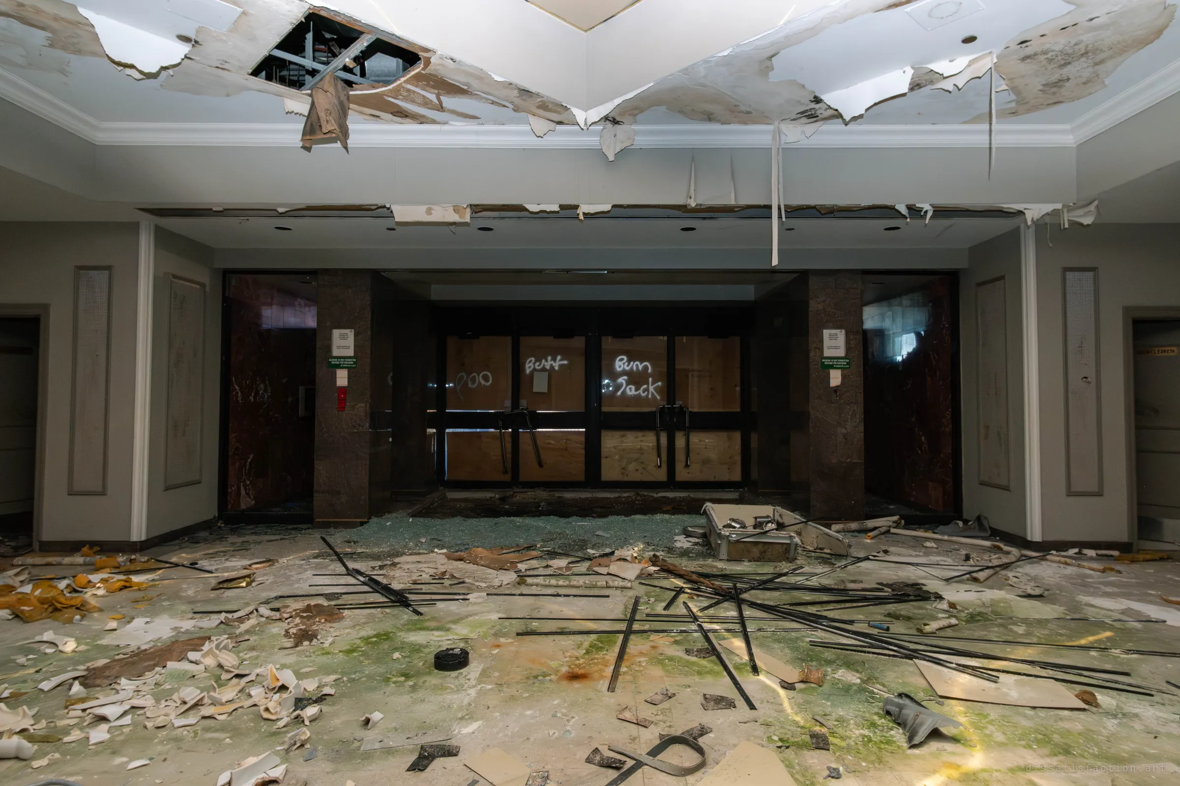 Boarded-up glass entrance doors with white spray-painted scrawls, flanked by dark marble pillars, with shattered glass, fallen ceiling plaster and scattered rods across the floor.