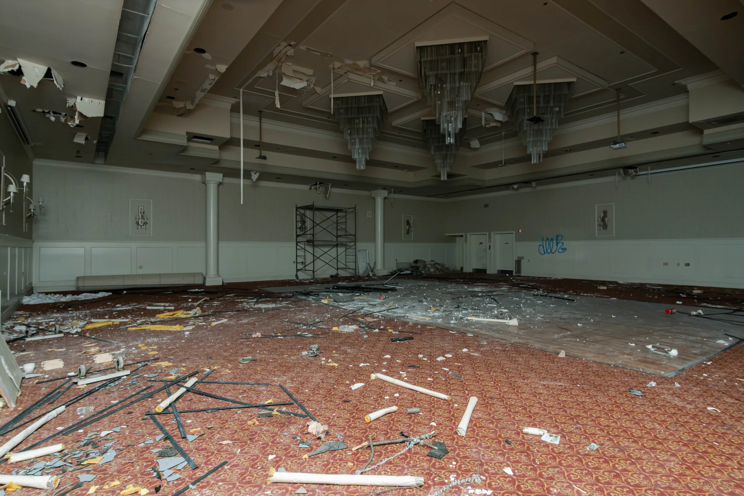 A ballroom with cluster chandeliers and a damaged ceiling, a scaffold tower against the far wall and a red patterned carpet with a bare patch, covered in rods and debris.