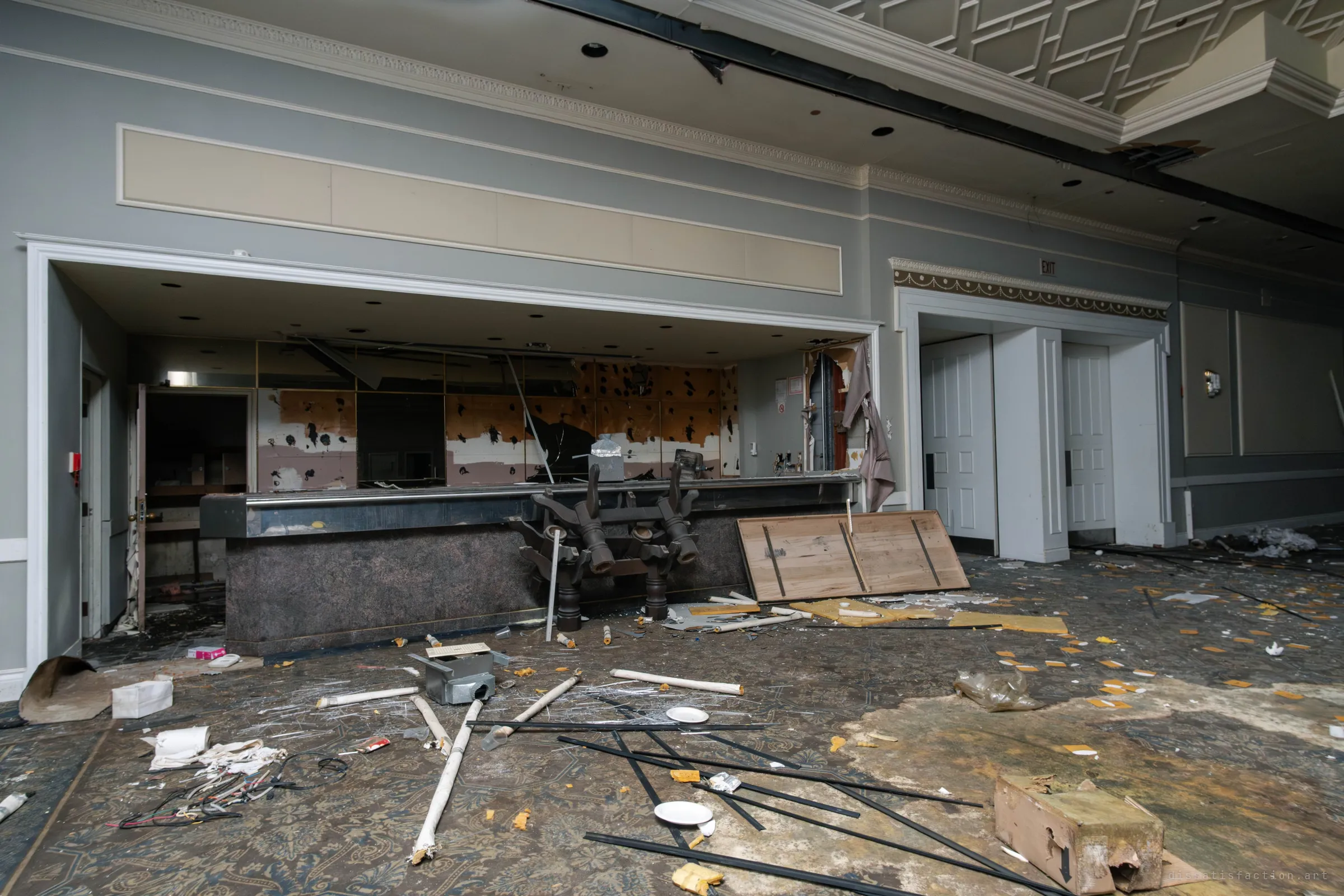 A bar opening and white double doors along the wall of a ballroom, with insulation, pipes and broken boards across the carpet and an upturned table base in front of the bar.