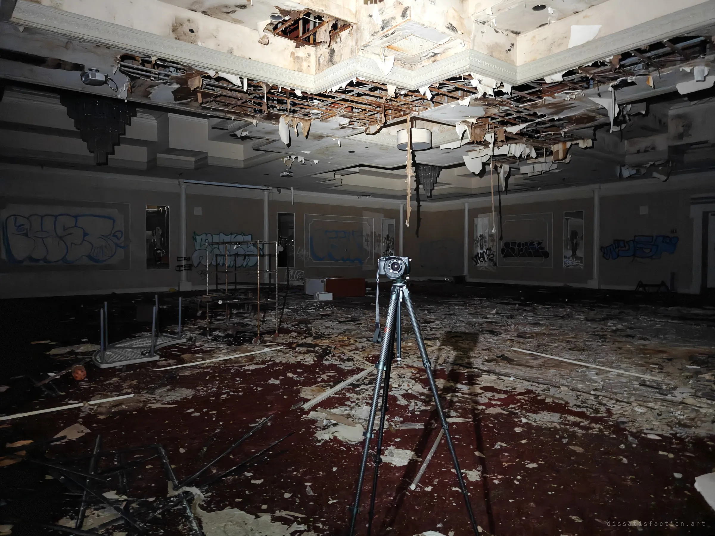 A wide ballroom with a collapsed, peeling ceiling and graffiti panels along the walls. A camera on a tripod stands in the middle of a debris-covered red carpet.