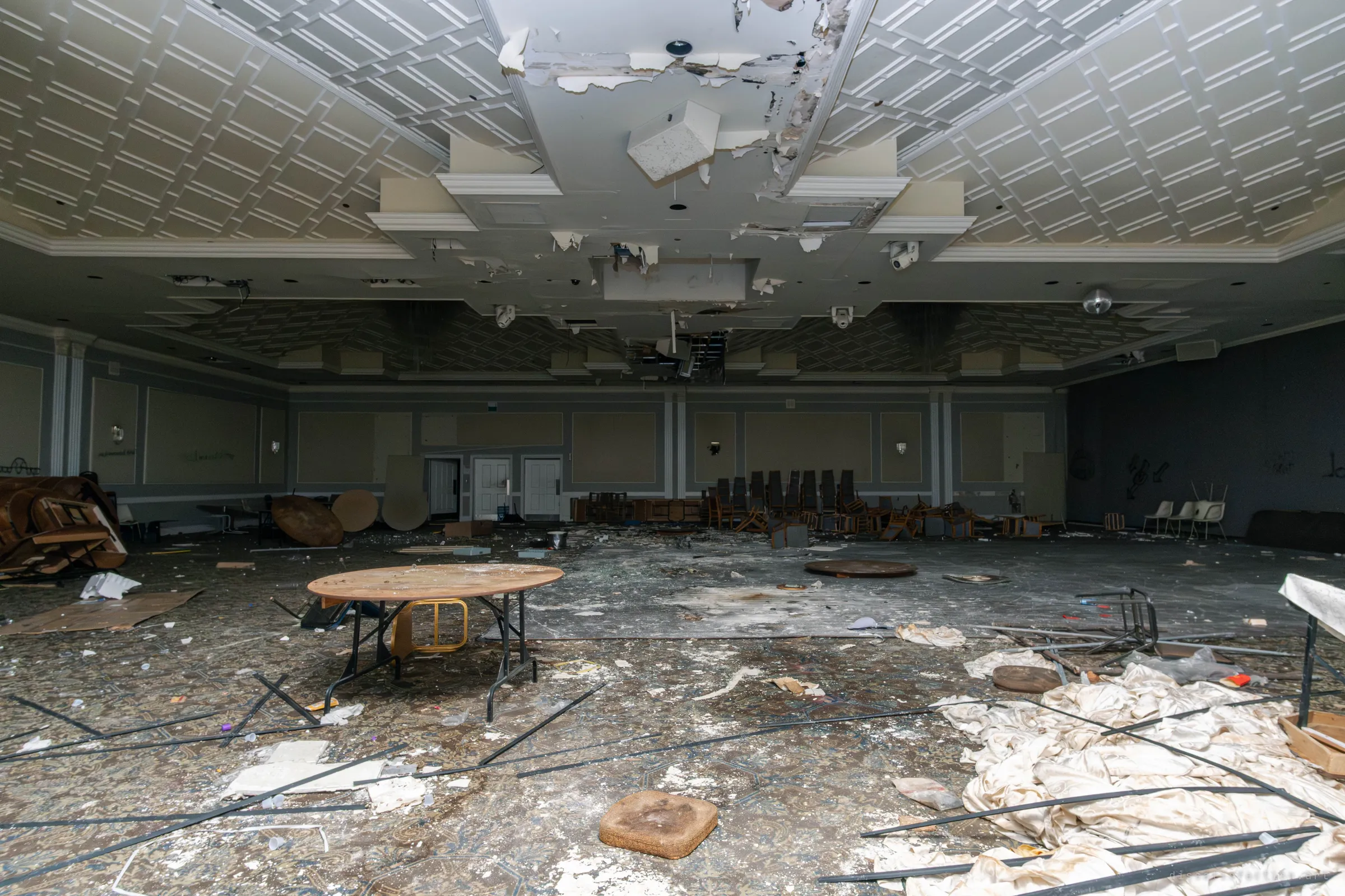 The same banquet hall seen from the opposite side, with a damaged ceiling, security cameras, a round table in the foreground and bedsheets piled on the filthy carpet.