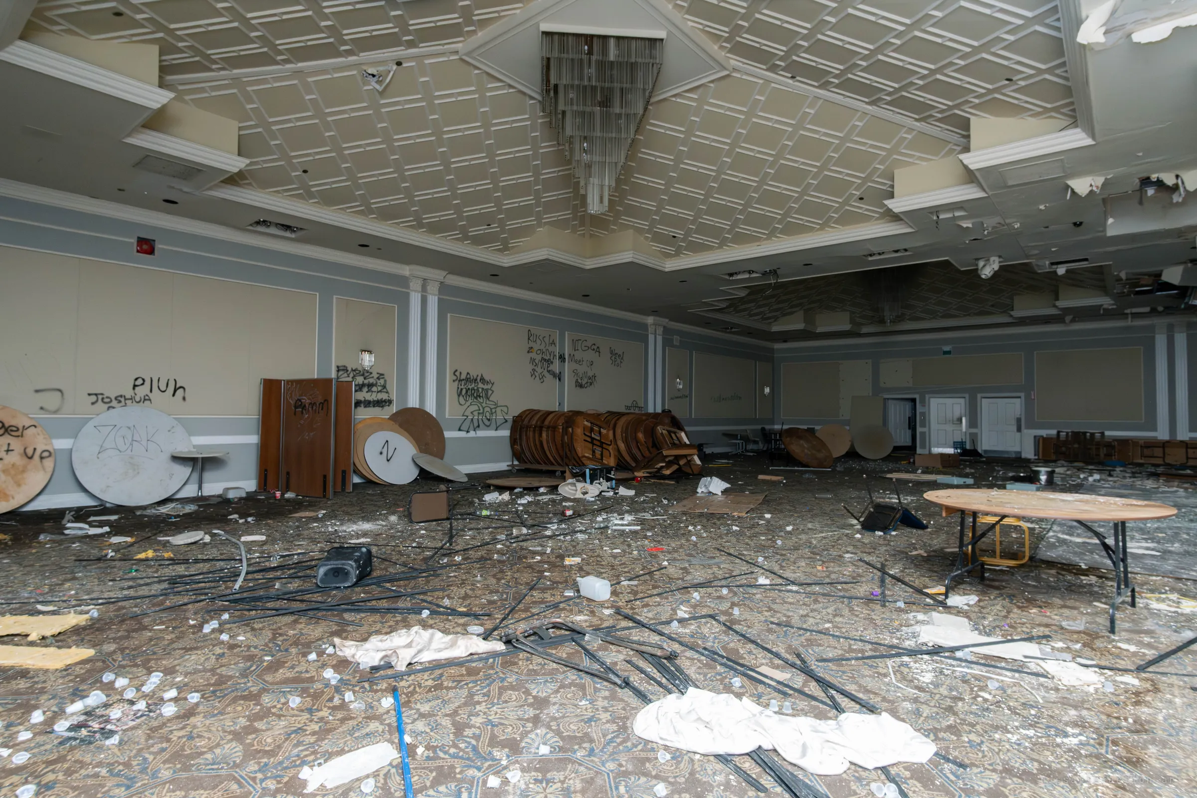 A large banquet hall with a coffered ceiling and a chandelier, with round tables stacked on their sides against graffiti-covered walls and debris covering a patterned carpet.