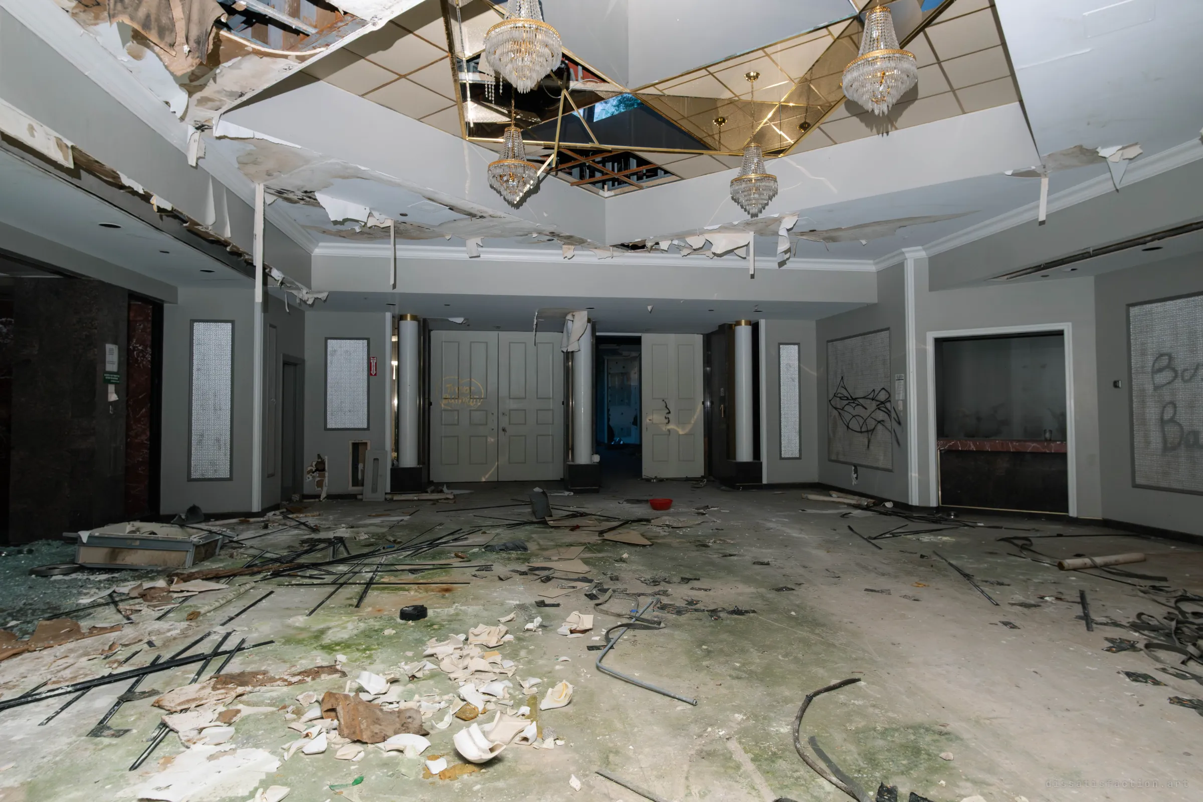 A wrecked entrance hall with a mirrored ceiling well, crystal chandeliers and peeling ceiling panels above a floor strewn with broken ceramic, black rods and debris, with tall white double doors at the far end.