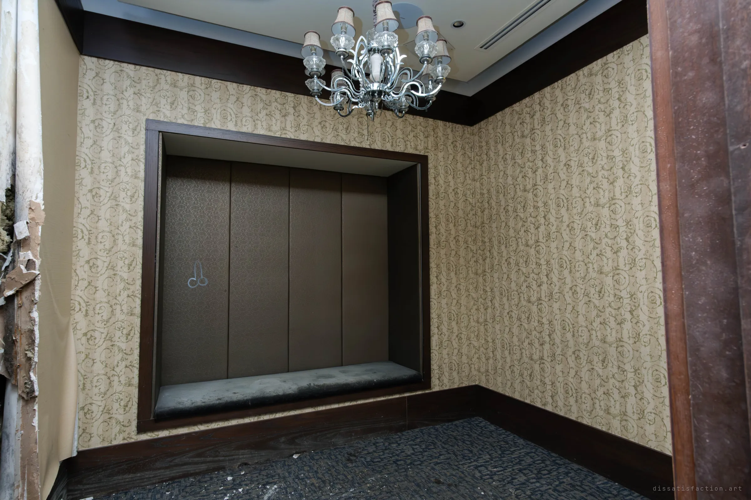 A small banquet side room with scrolled wallpaper, a padded bench alcove and a chandelier, still intact. A faint chalk drawing sits on one of the panels, and peeling wall at left shows the decay.