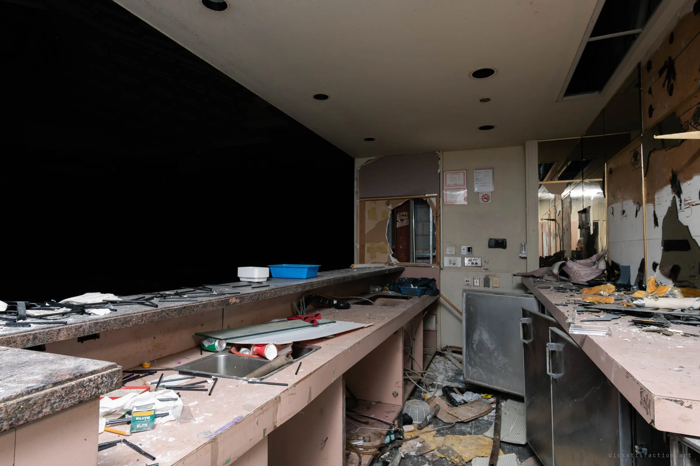 A gutted back-of-house kitchen with pink laminate counters and sinks, a stainless steel cabinet, smashed wall mirrors and a cigarette pack and debris on the work surface, with a dark void to the left.