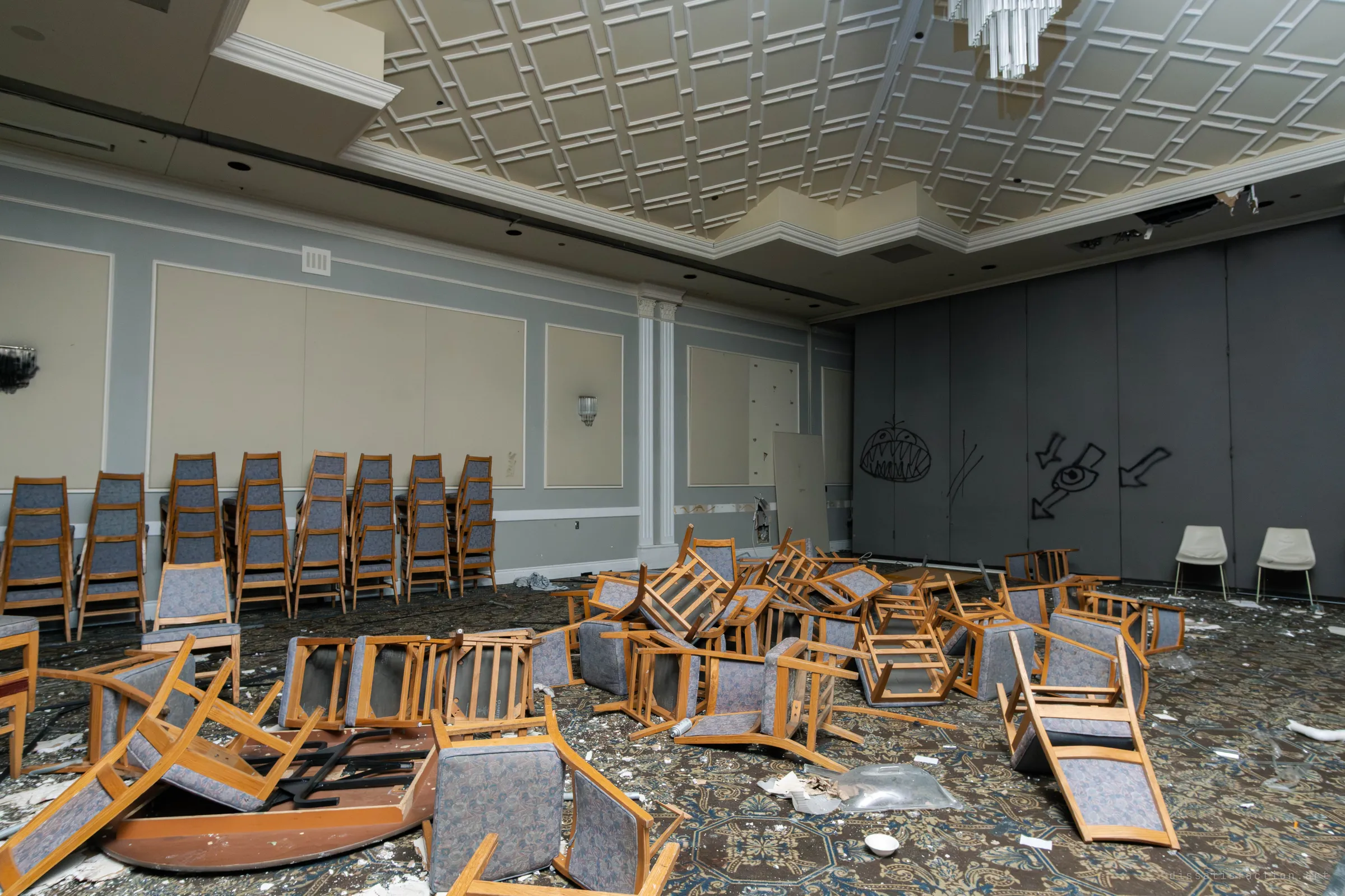 Stacked blue upholstered banquet chairs against a pale wall beside a heap of overturned chairs, with a grey folding partition wall covered in black graffiti monsters and arrows.