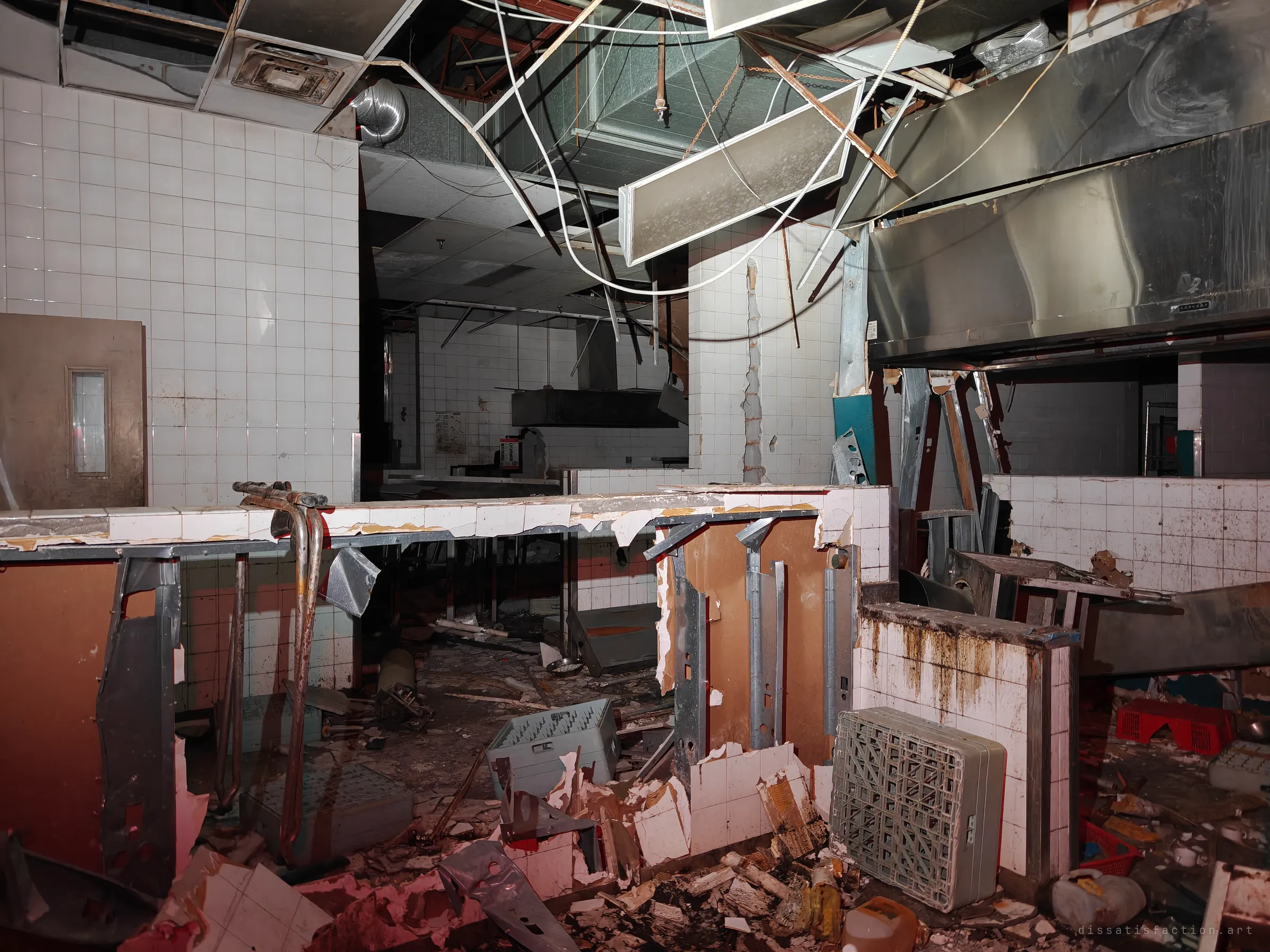 A gutted commercial kitchen where white-tiled walls and counters have been smashed open. Cables hang from a ceiling that has fallen onto crates and rubble, under a red-tinted light.