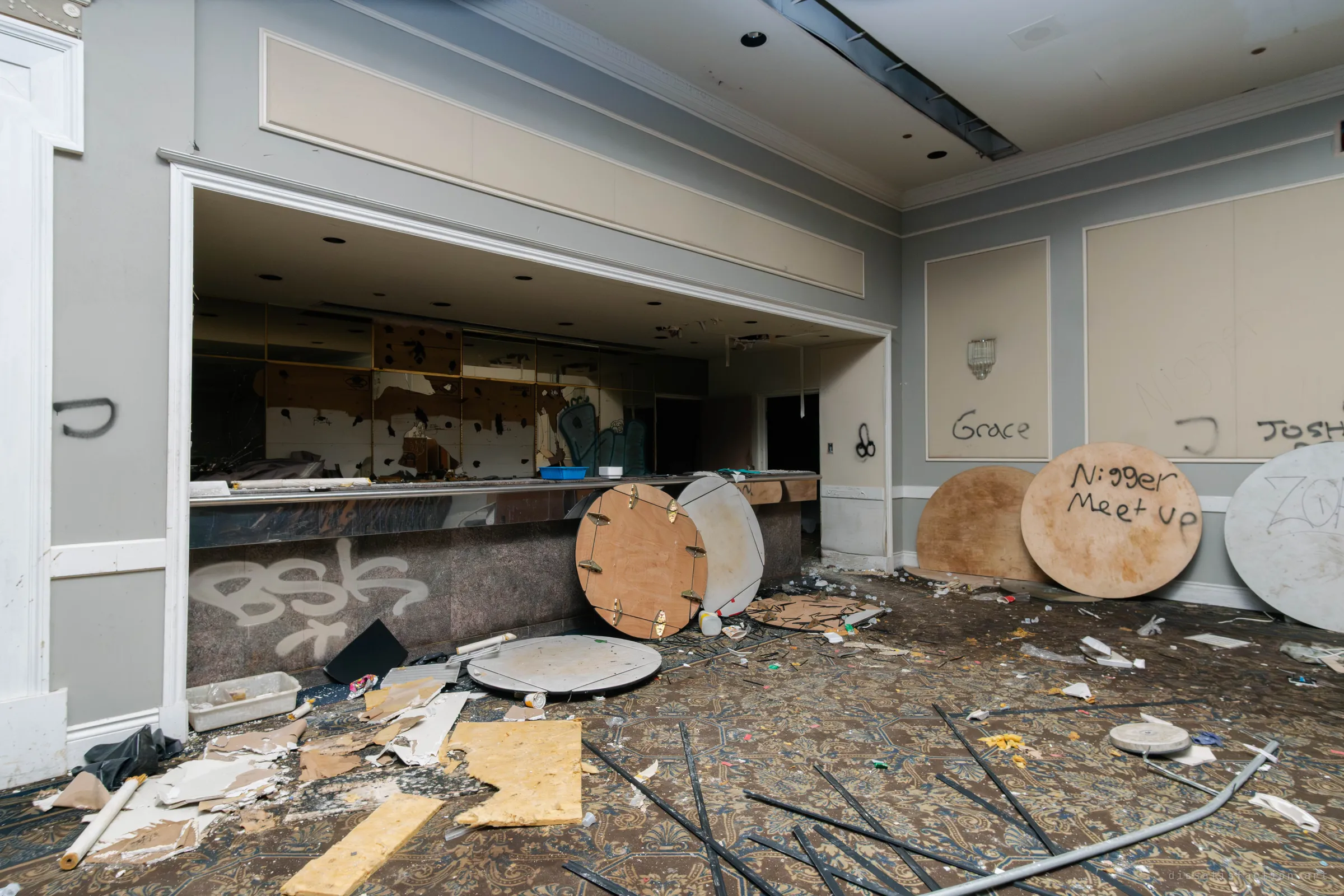 A serving bar opening in a banquet hall with a tagged granite counter, upended round table tops leaning on it and torn plaster and insulation scattered over the carpet.