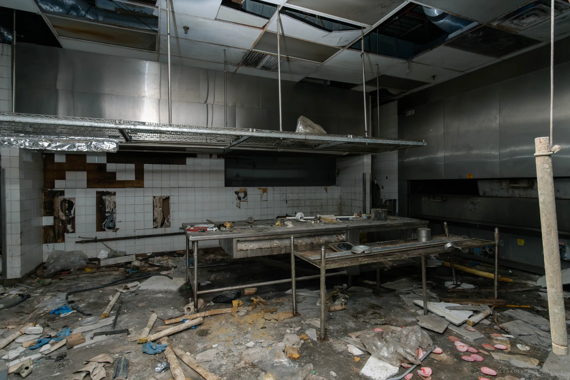 A ruined commercial kitchen with a stainless steel prep table, a wall of white tiles torn out in patches and a sagging ceiling. The floor is covered in blue gloves, pipes and plastic.