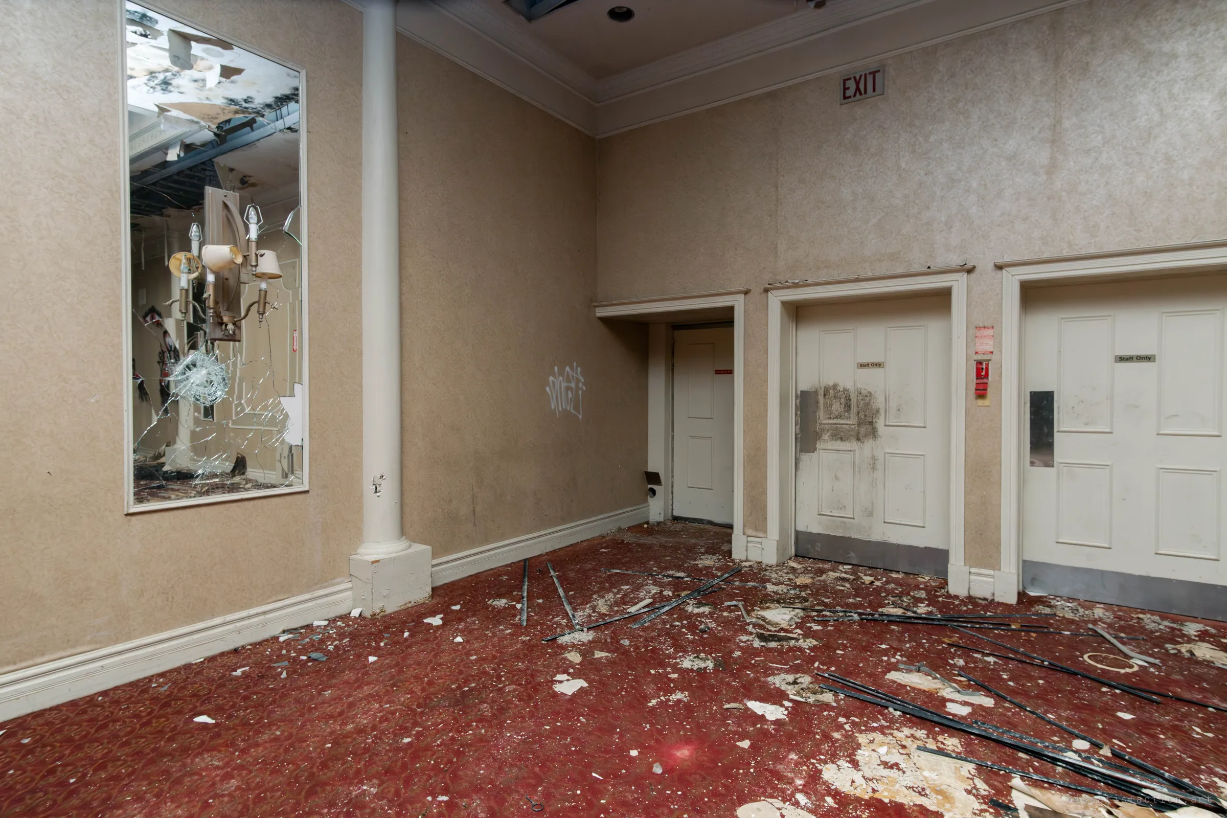 A corner by white staff-only doors with a shattered wall mirror reflecting a chandelier and collapsed ceiling, above a red carpet scattered with plaster and rods.
