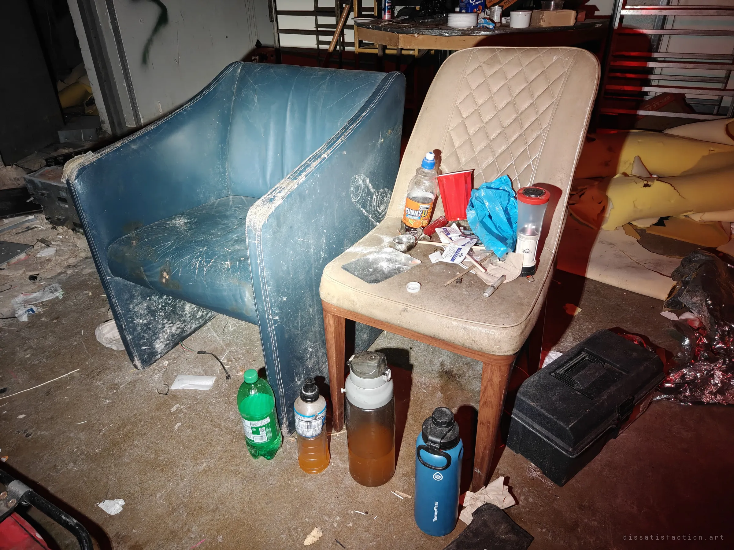 A worn blue armchair beside a cream chair covered in bottles, a plastic cup and small packets. Water bottles and a toolbox stand on the floor, evidence of someone sheltering in the building.
