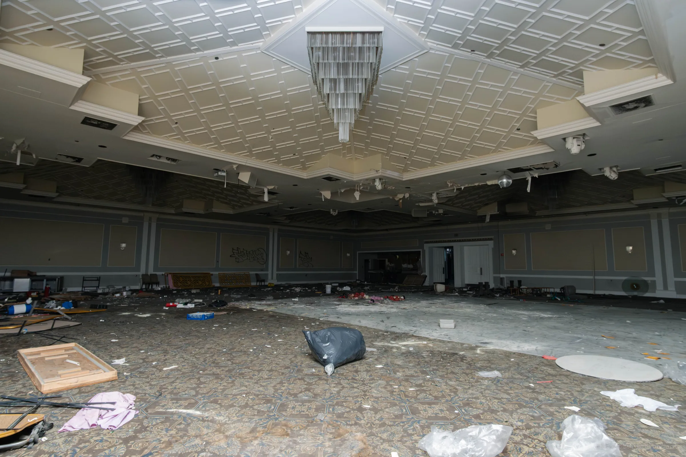 A wide banquet hall under a coffered ceiling with a crystal chandelier, hanging ceiling lights and security domes above a patterned carpet scattered with trash bags and fabric.