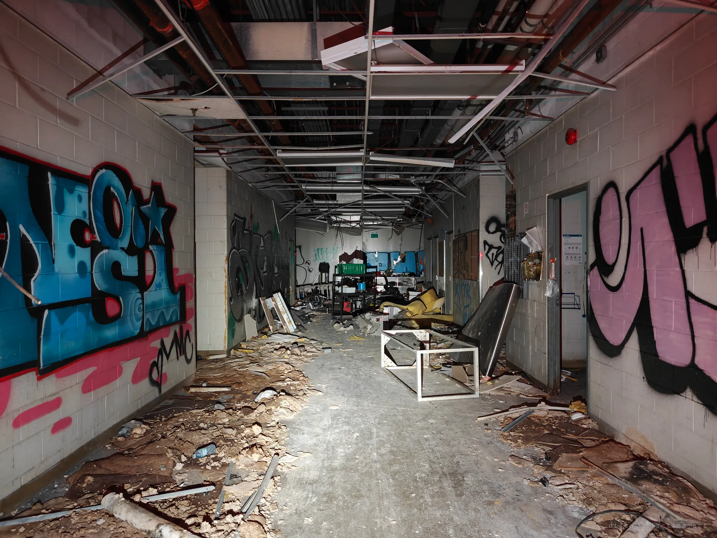 A service corridor with bare ceiling grid hanging overhead and huge blue and pink graffiti pieces along the block walls. Wrecked furniture and debris line the way to a room at the far end.
