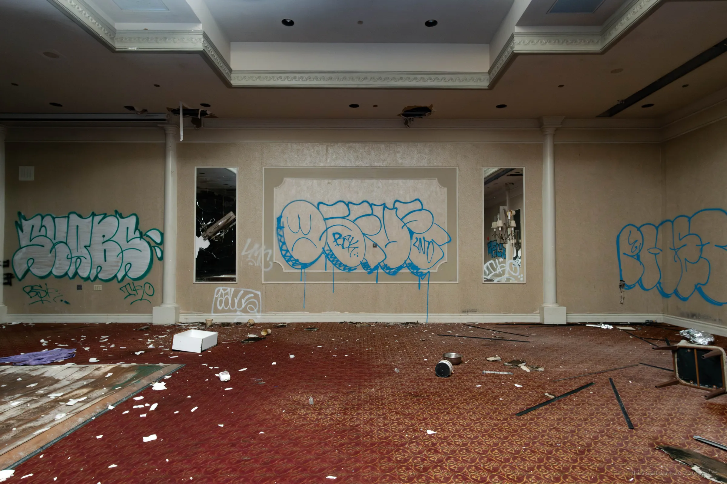 A room with mirrored panels and large blue and teal graffiti pieces on beige walls, with a red patterned carpet strewn with plaster shards and a patch of bare wooden dance floor at left.
