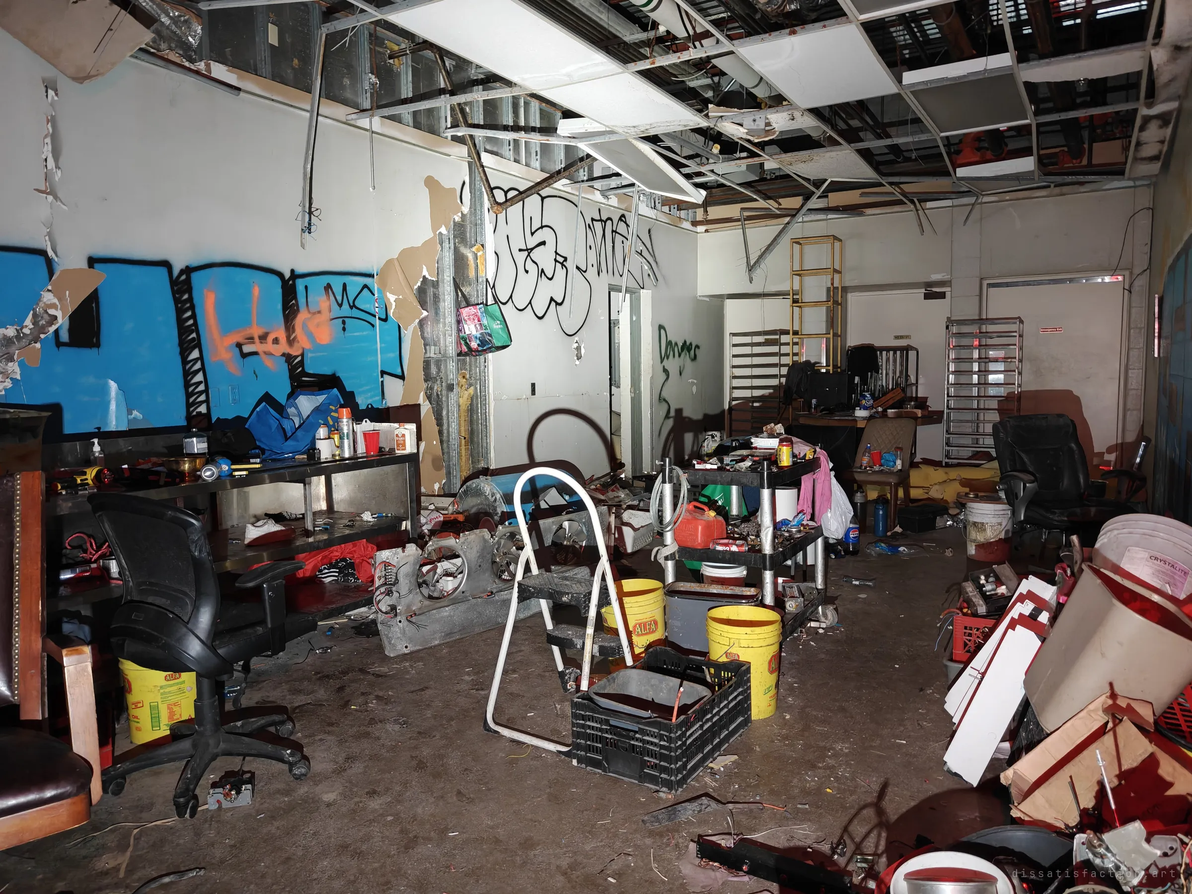 A cluttered back room where someone has been camping, with office chairs, buckets, a stepladder and shelves of tools. A large blue and orange graffiti piece covers the wall behind them.