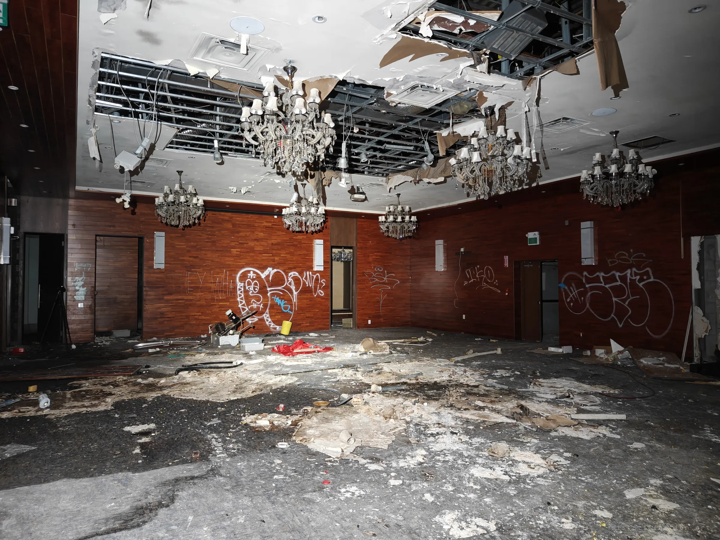 A main banquet hall with red wood walls, five chandeliers and a ceiling peeled open to its metal frame. Graffiti tags cover the walls above a floor thick with plaster.