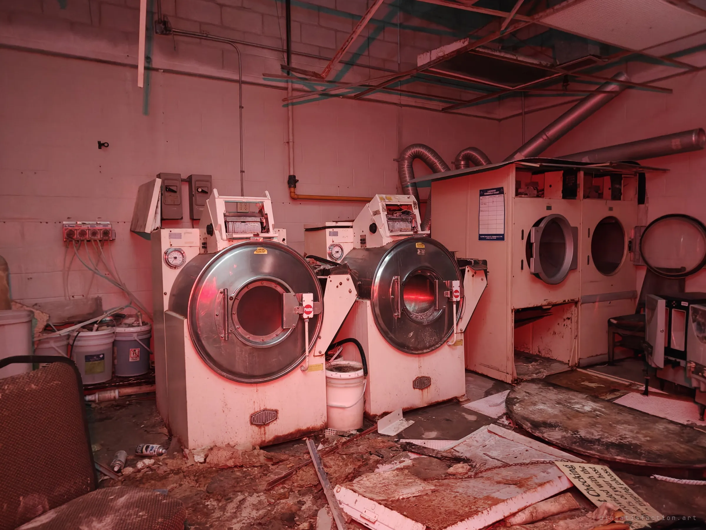 Two industrial washing machines and a double dryer in a rusted laundry room, lit in red. Detergent buckets, ducting and a fallen sign lie among the debris.