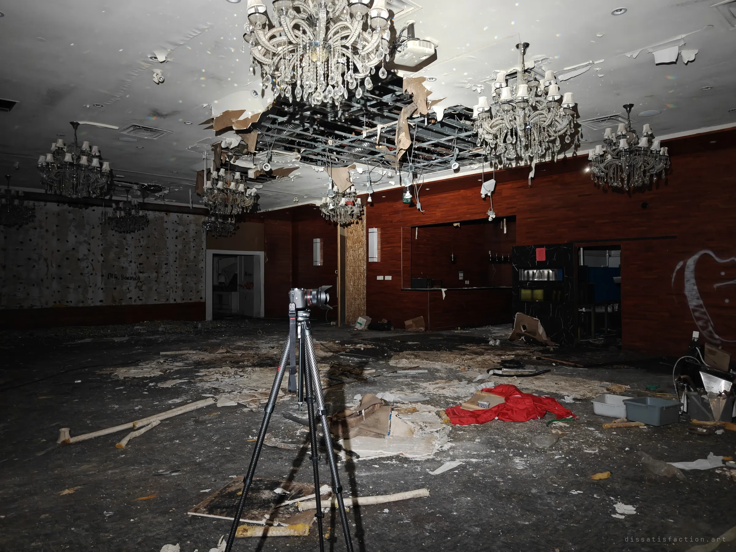 A banquet room with crystal chandeliers, a half-fallen ceiling and dark wood paneling. A tripod stands on a floor of plaster flakes, with a red cloth lying near the far wall.